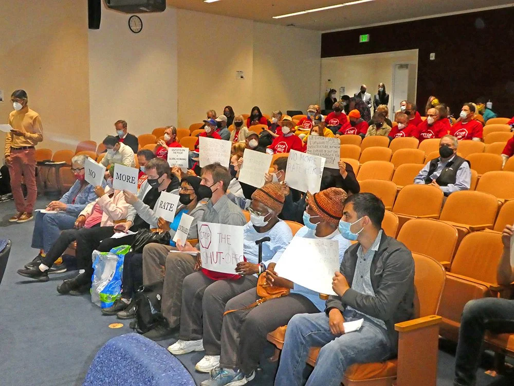 A mixed group of people filling the front rows of a hearing room with signs that say, "NO MORE RATE INCREASES" and "STOP THE SHUT OFFs"
