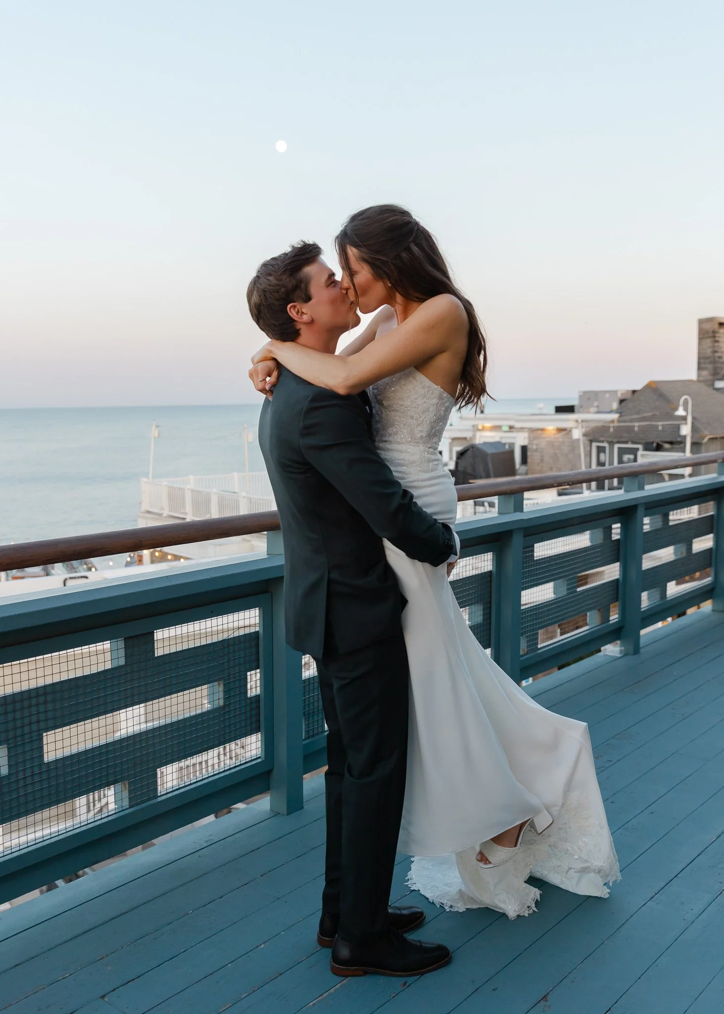 A bride and groom sharing a kiss on a balcony with ocean view during sunset.