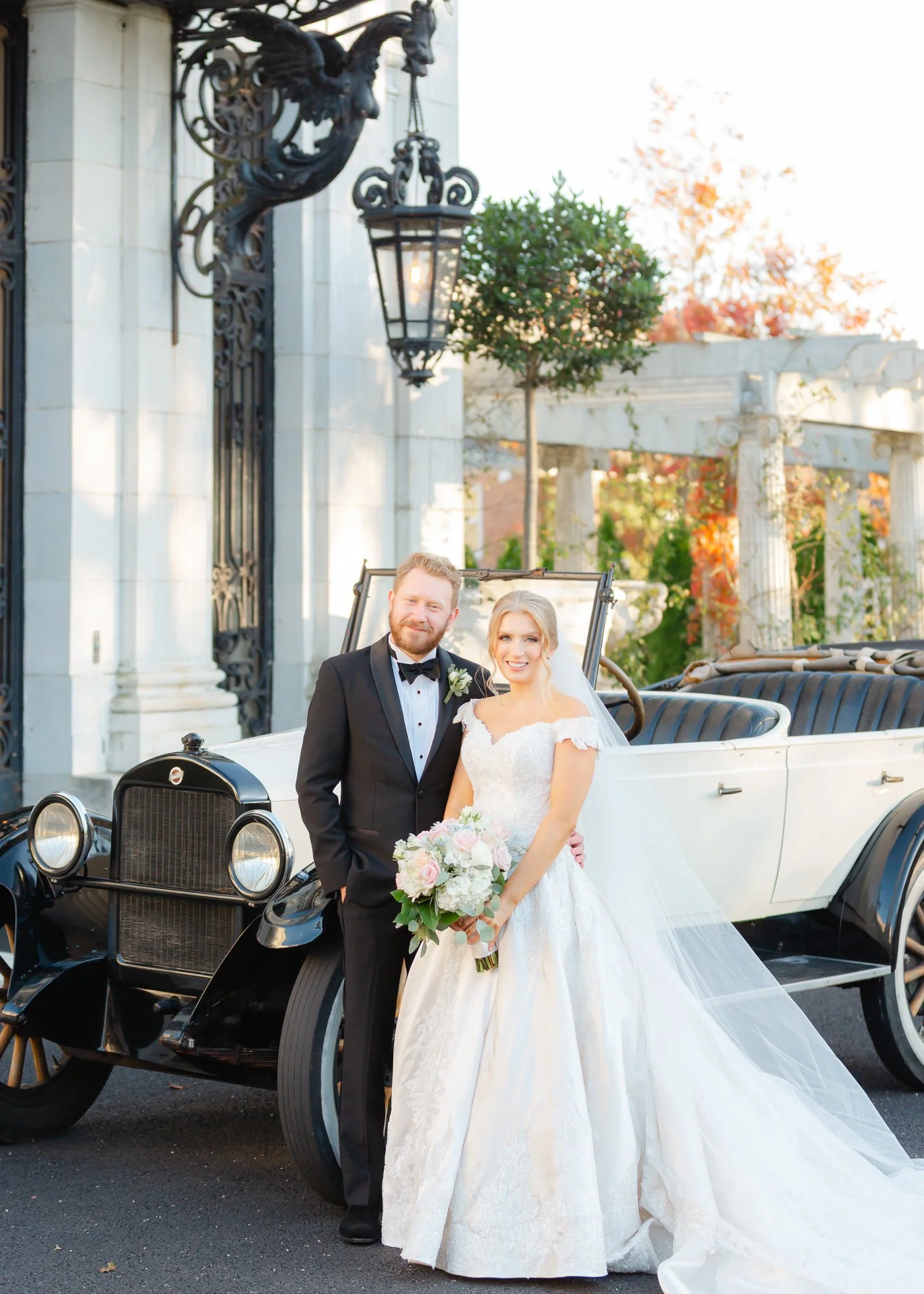 A newlywed couple, a groom in a black tuxedo and a bride in a white wedding gown, standing in front of a vintage white and black car, with an ornate gate, lantern, and columns in the background.