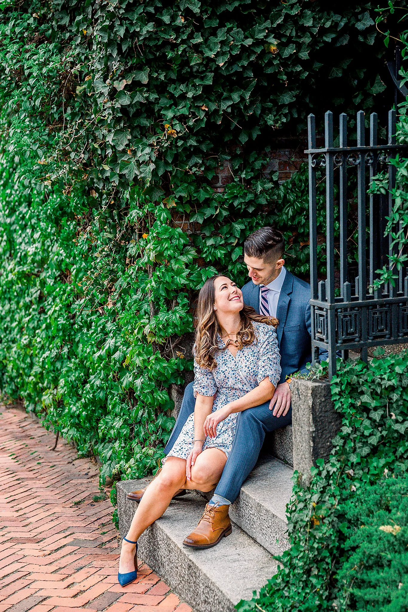 A woman and a man sitting on a stone bench surrounded by lush green ivy, smiling at each other.