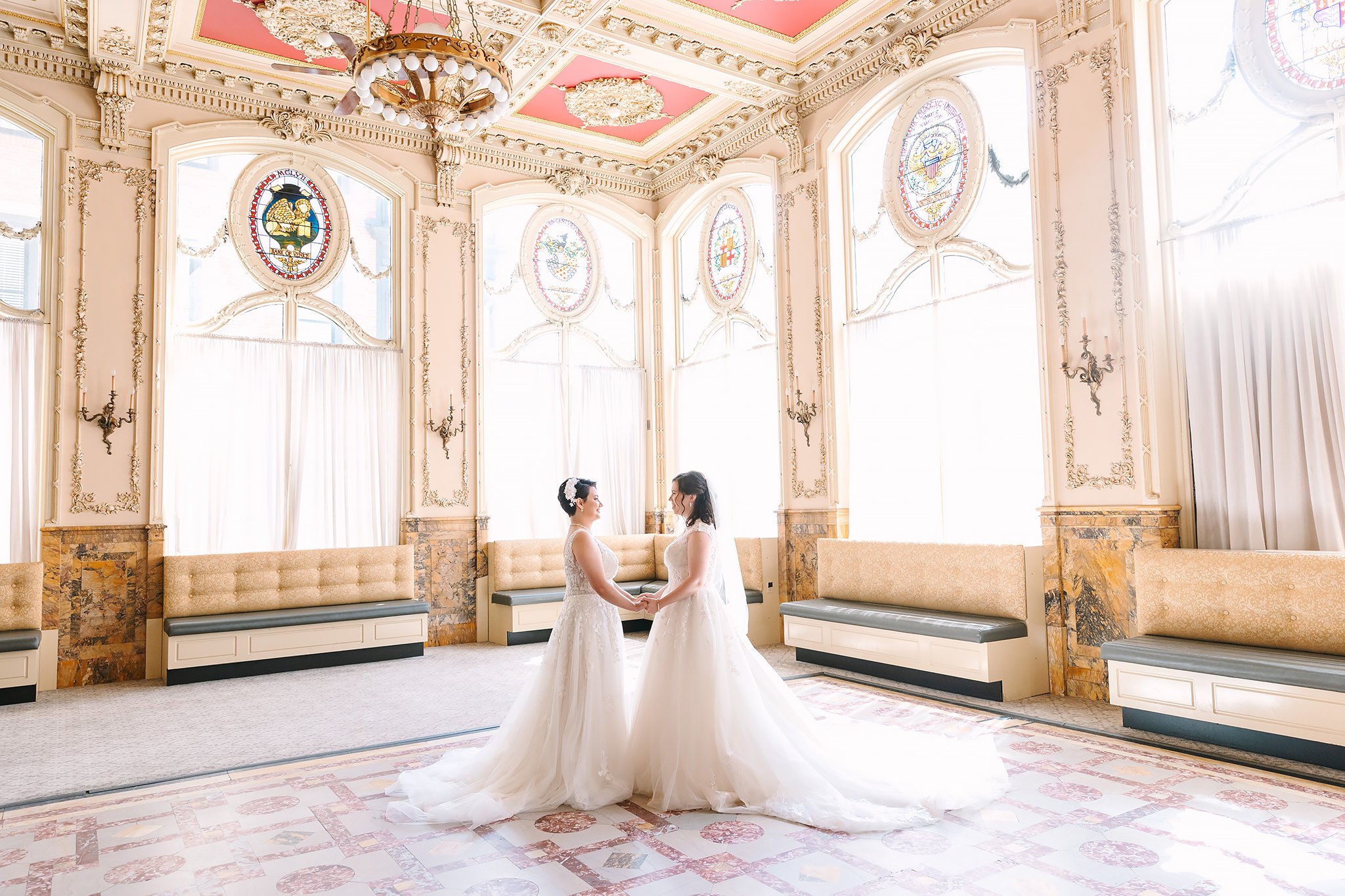 Two brides in wedding dresses holding hands in a bright, ornate room with large stained glass windows and elegant decor.