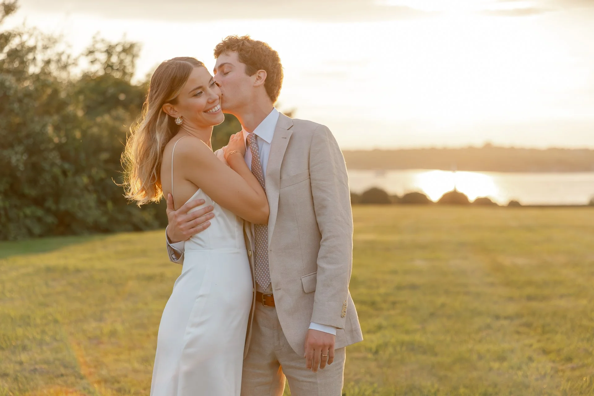 A couple dressed in wedding attire embracing outdoors at sunset by a body of water.