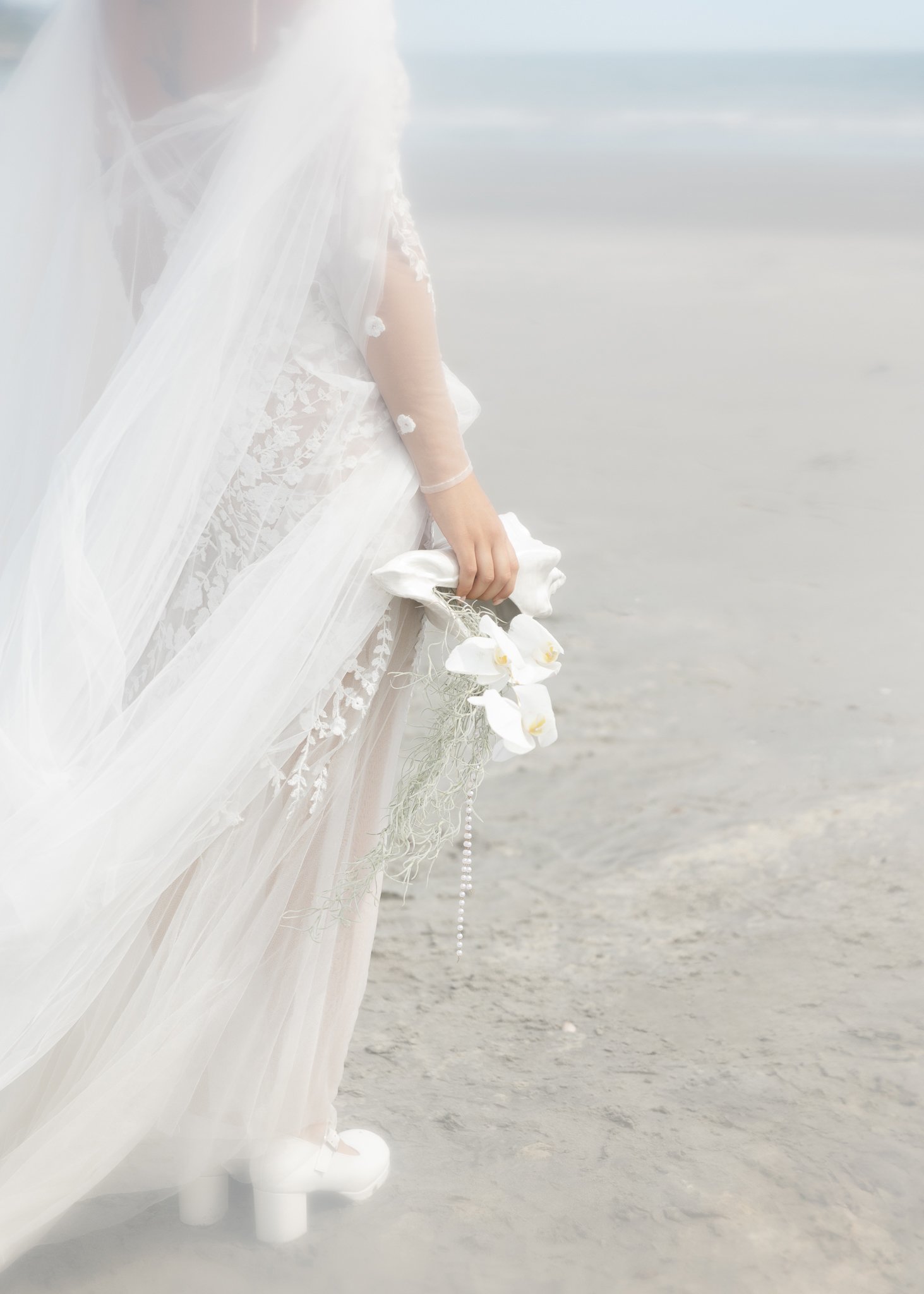 Close-up of a bride in a white wedding dress and veil holding a bouquet of white orchids on a beach.