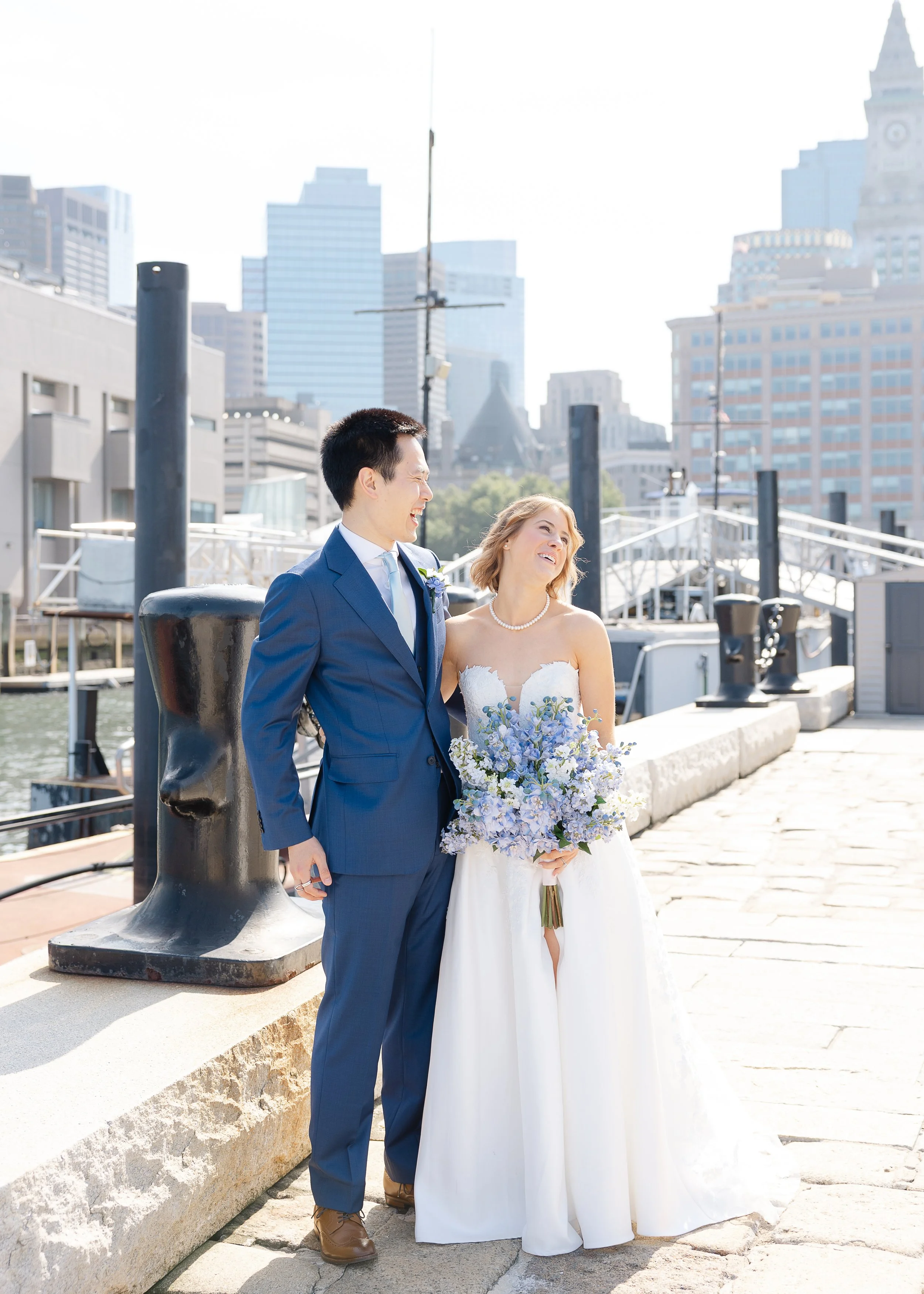 A bride and groom stand together outdoors at a marina with city buildings in the background, smiling and looking at each other, with the bride holding a bouquet of blue and white flowers.