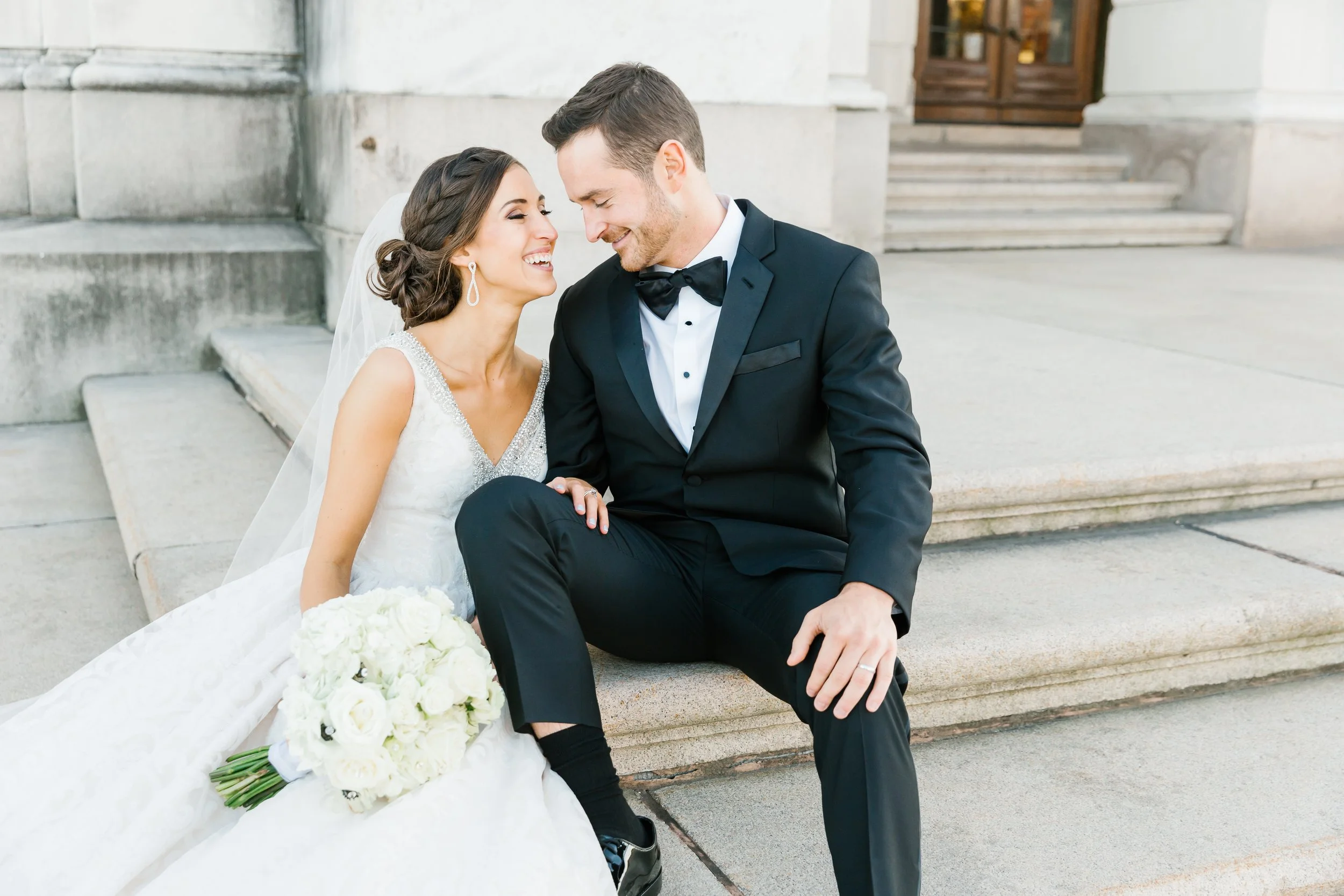 A newlywed couple sitting on steps outside, smiling and leaning their foreheads together. The bride is in a white wedding dress holding a bouquet of white roses, and the groom is in a black tuxedo with a bow tie.