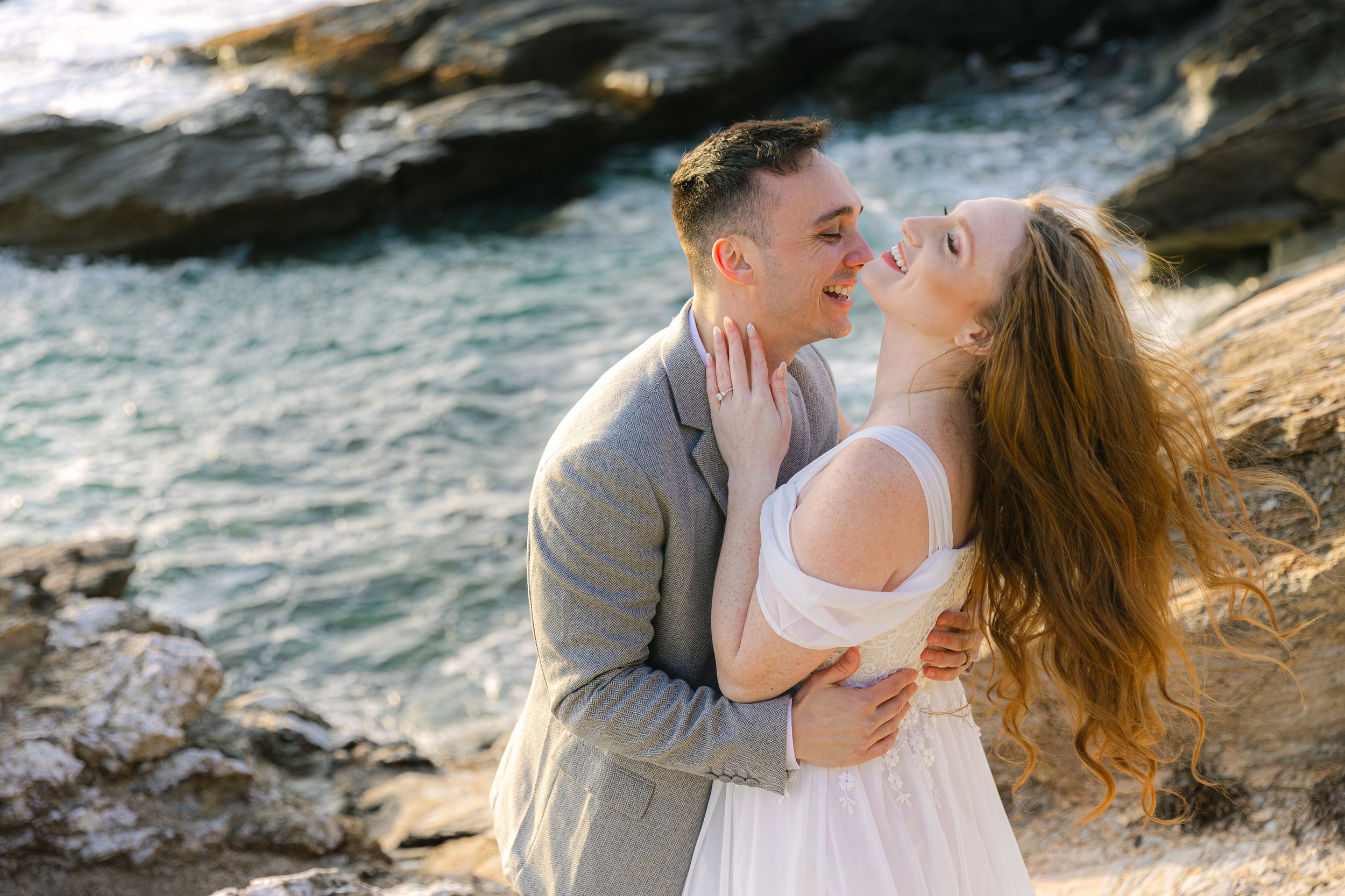 A smiling couple embraces on a rocky shoreline with water behind them. The woman has long red hair and is wearing a white dress, and the man is in a light gray suit.