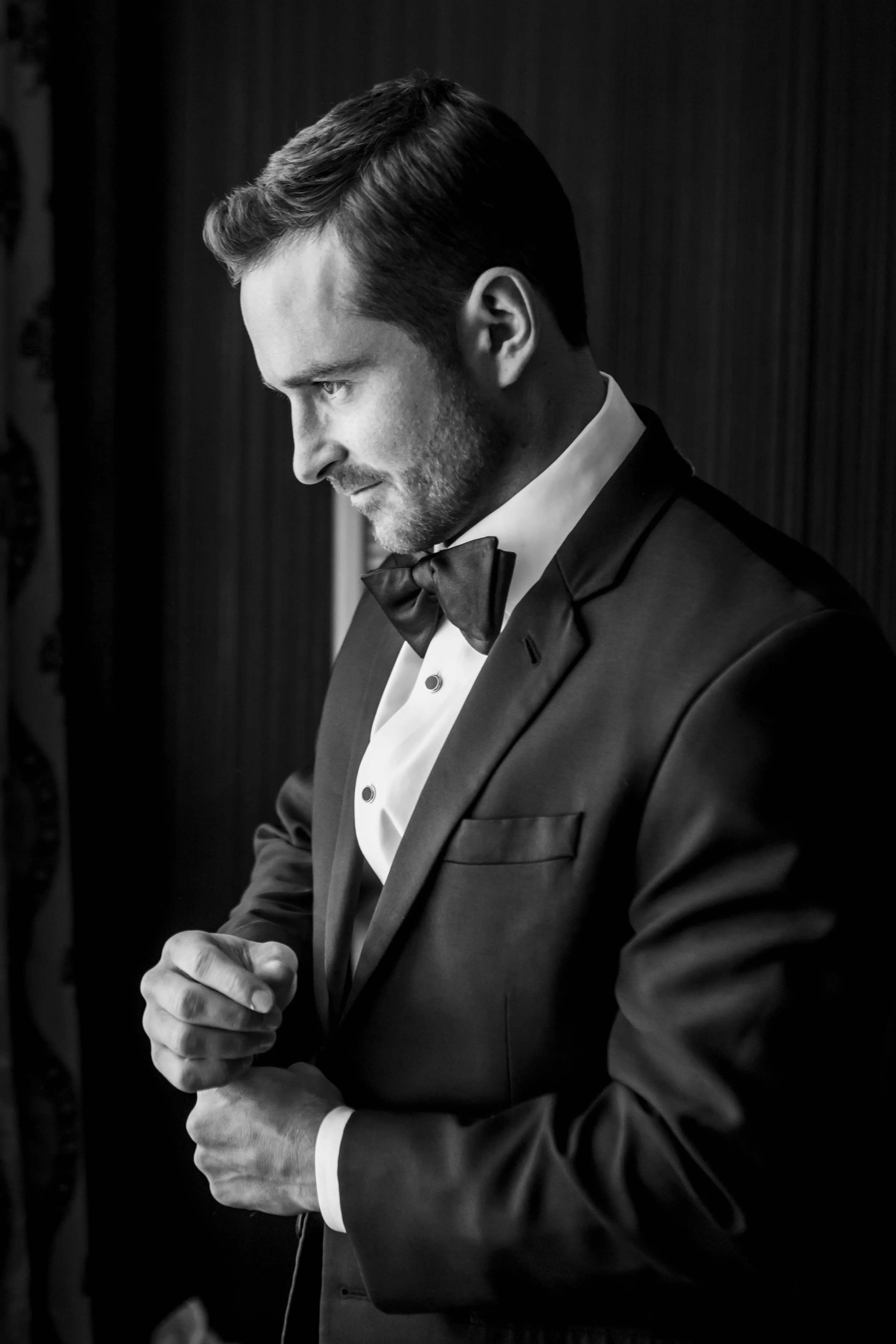 A man in a tuxedo and bow tie, adjusting his cufflinks, with a serious expression, in a black-and-white photograph.