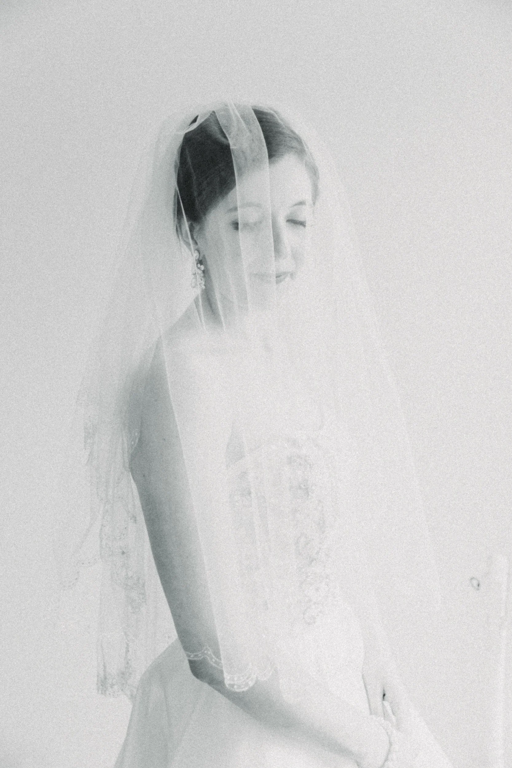 A black and white photo of a bride with closed eyes, wearing a veil and earrings, with her head slightly bowed.