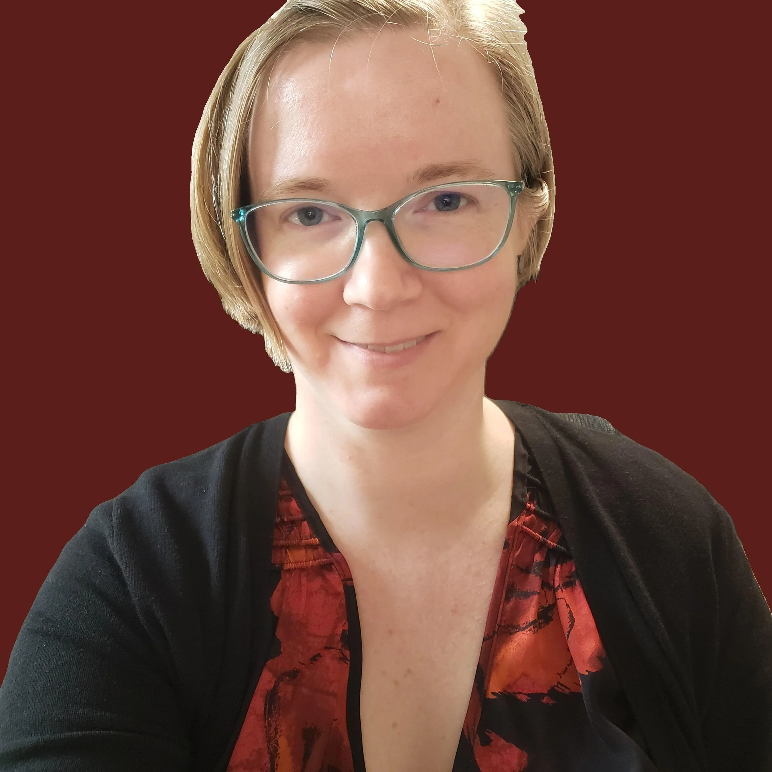 A woman with short strawberry blonde hair, wearing glasses, a patterned orange and black top, and a black cardigan, smiling in front of a solid dark red background.