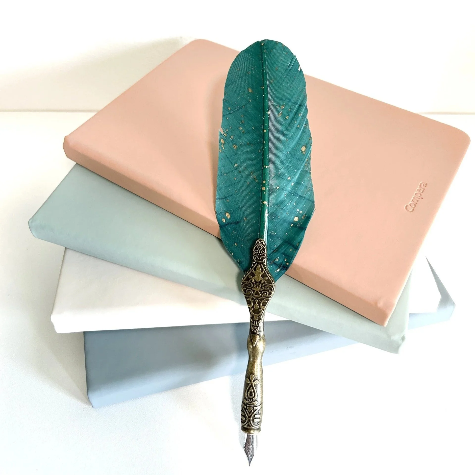 A teal decorative feather with ornate tarnished gold handle rests on four pastel-colored books in peach, sage, white, and light blue.