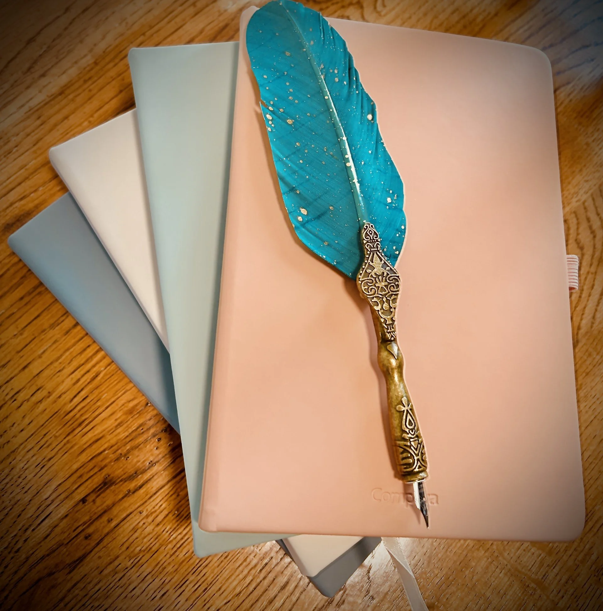 A decorative teal feather quill pen resting on closed pastel-colored notebooks on a wooden surface.