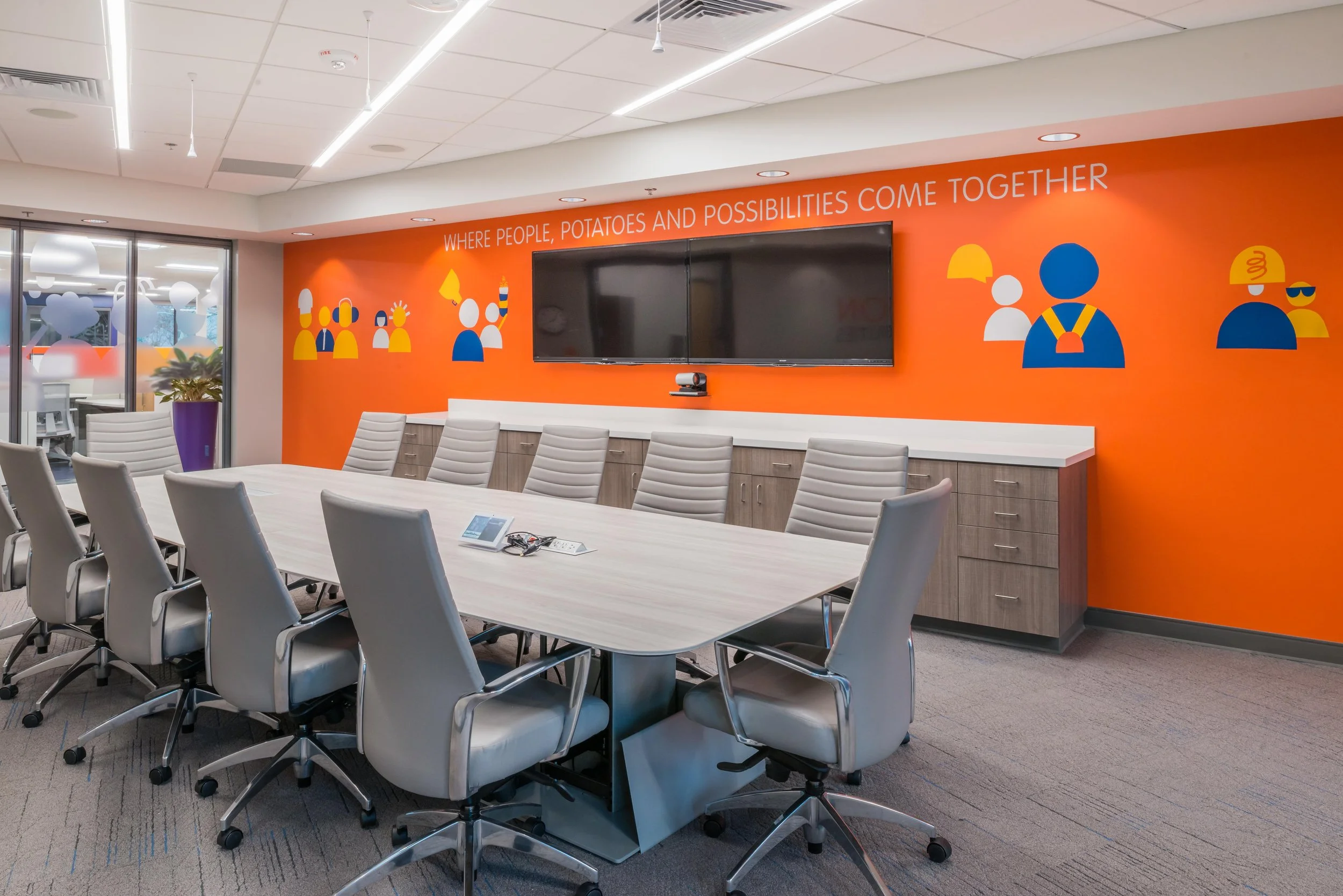 A modern conference room with a large table and multiple gray office chairs. The room features an orange accent wall with colorful graphic illustrations and the quote 'WHERE PEOPLE, POTATOES AND POSSIBILITIES COME together.' There is a large flat-screen TV on the wall and sliding glass doors leading to an outdoor patio area.
