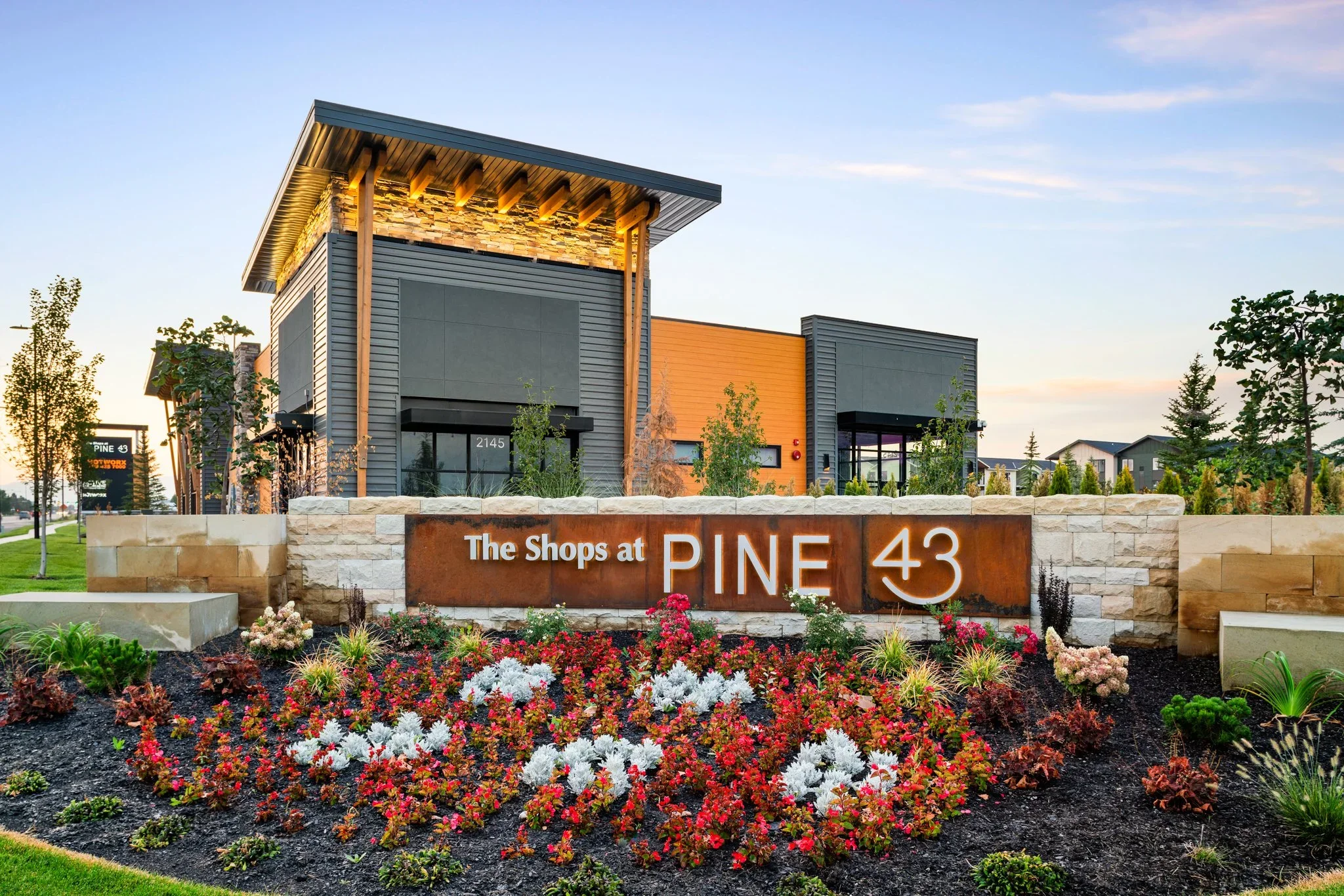 Entrance sign for The Shops at Pine 43 with colorful flower bed in front and modern building in background during sunset.