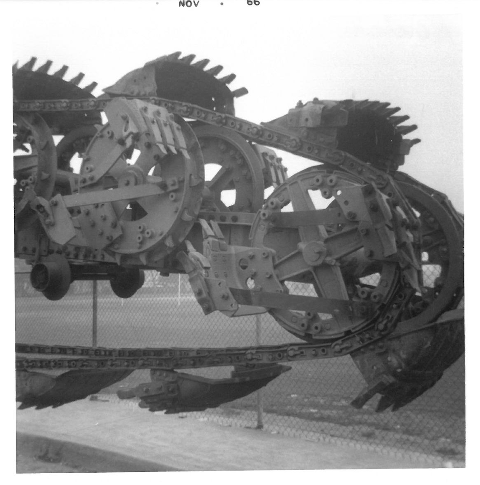 Close-up of a large metal sculpture resembling a fish, constructed from mechanical parts, on display outside in front of a chain-link fence.