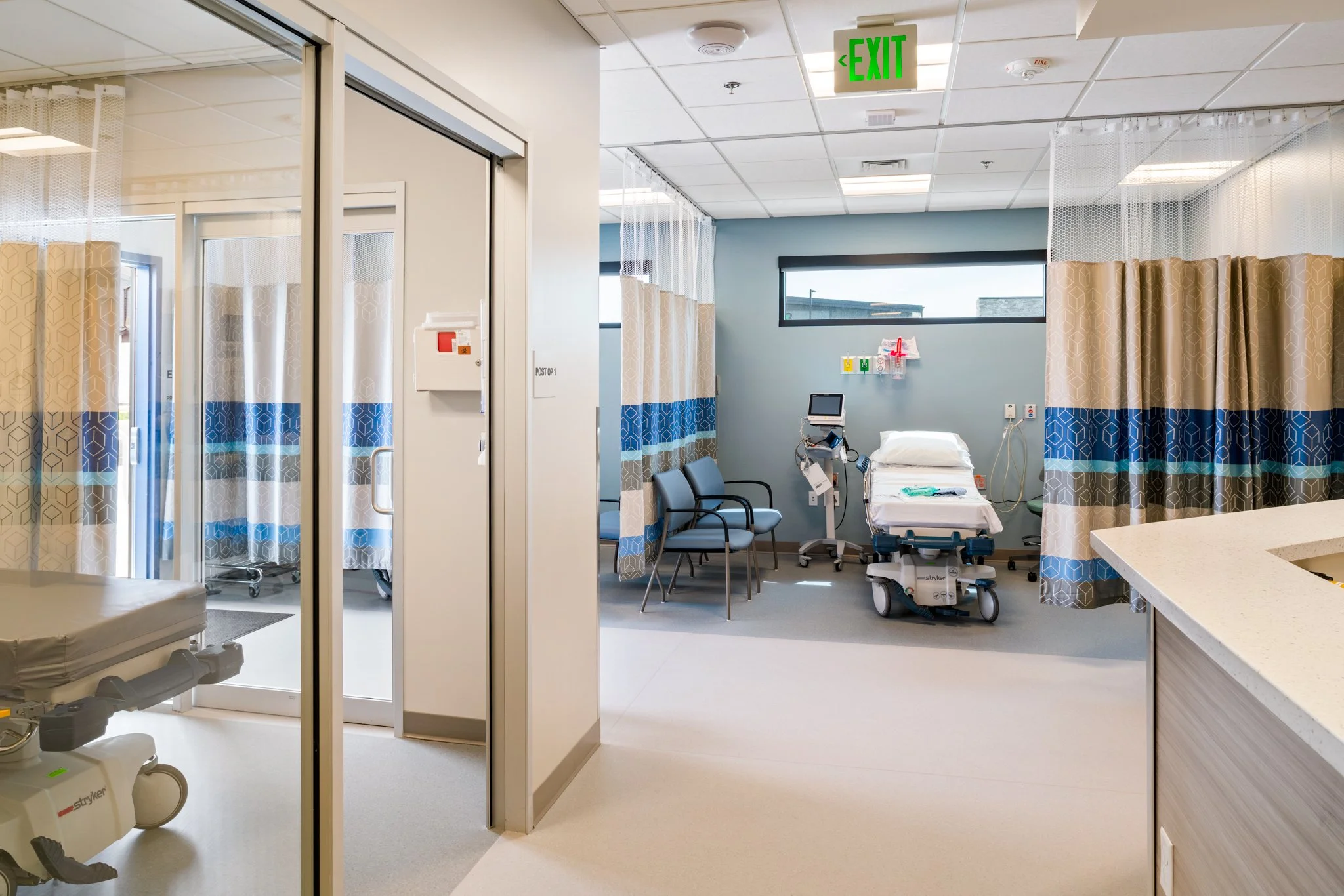 Hospital room with medical equipment, a hospital bed, chairs, privacy curtains, and an illuminated green exit sign on the ceiling.
