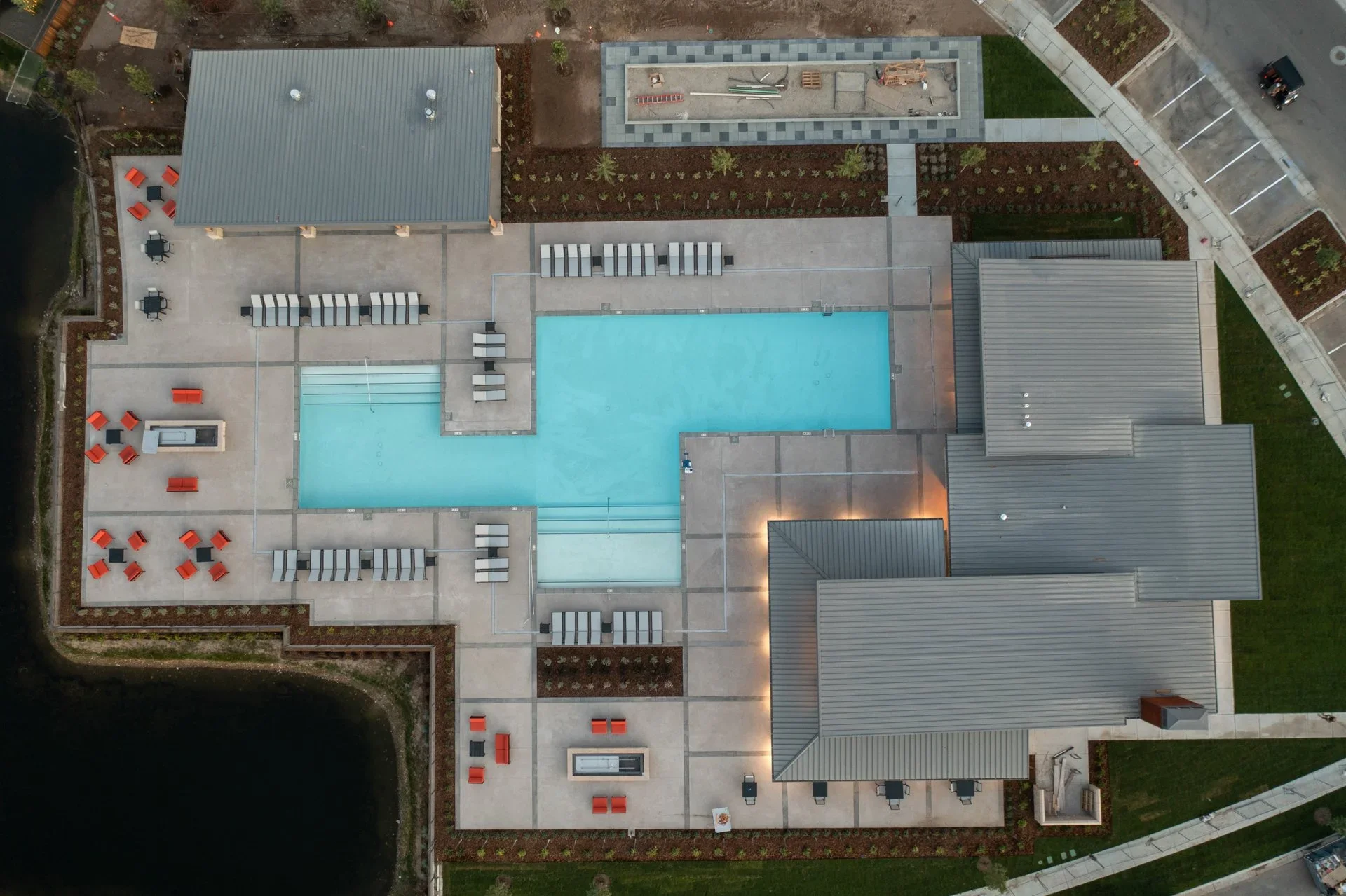 Aerial view of a modern outdoor swimming pool area with lounge chairs, tables, and umbrellas, surrounded by a landscaped garden, parking lot, and adjacent buildings.