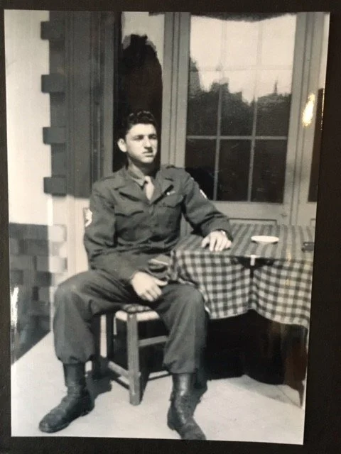 Black and white photo of a man in military uniform sitting on a chair with one hand on a table and outdoors behind a window.