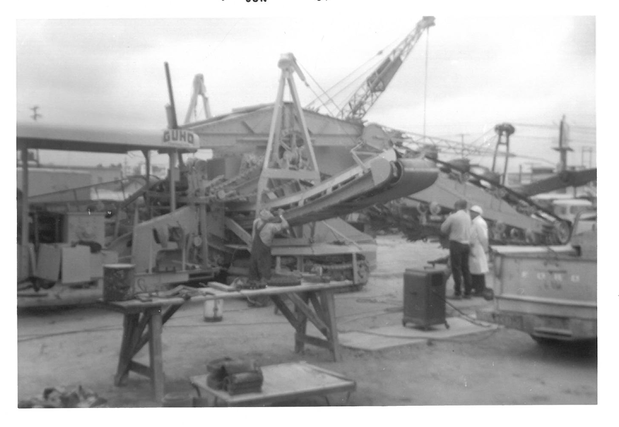 Black and white photograph of a work area with several large cranes, military tanks, and two people wearing helmets and work clothes standing near equipment.