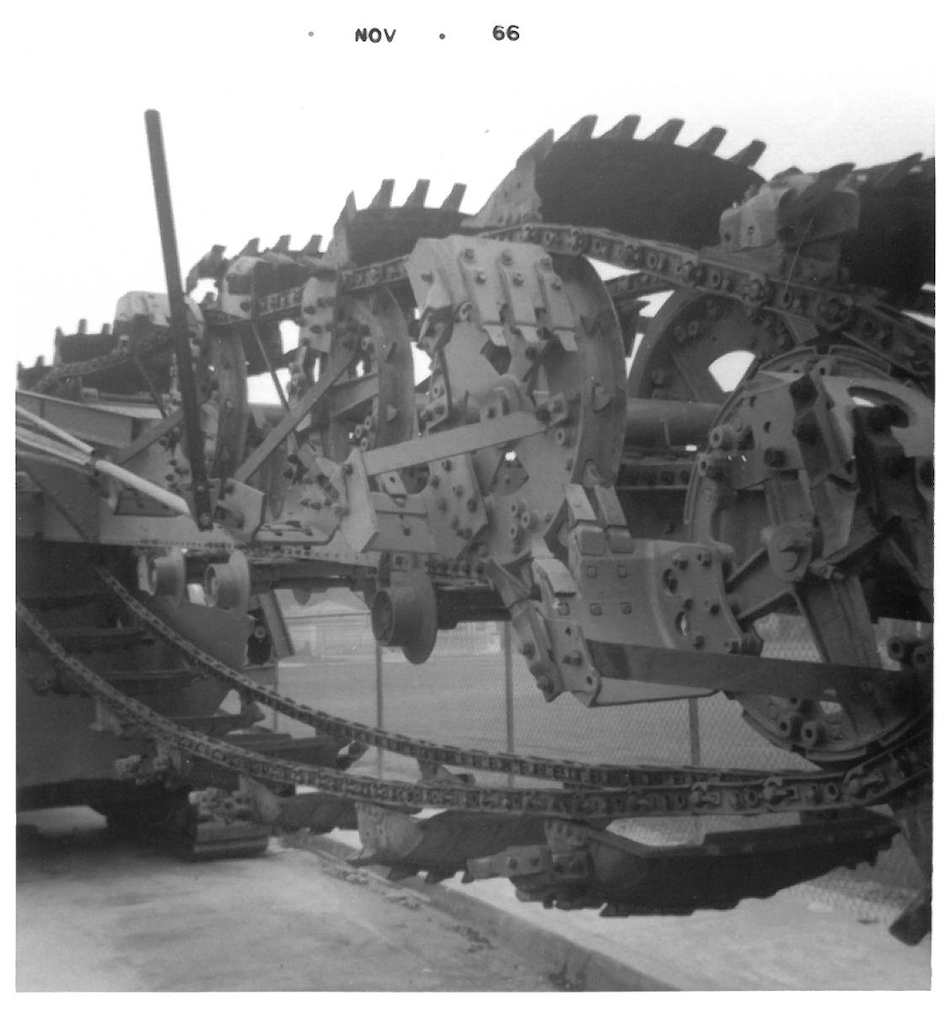 A black and white photograph of a large mechanical structure, possibly a tank or construction equipment, showing intricate gears, chains, and metal components.