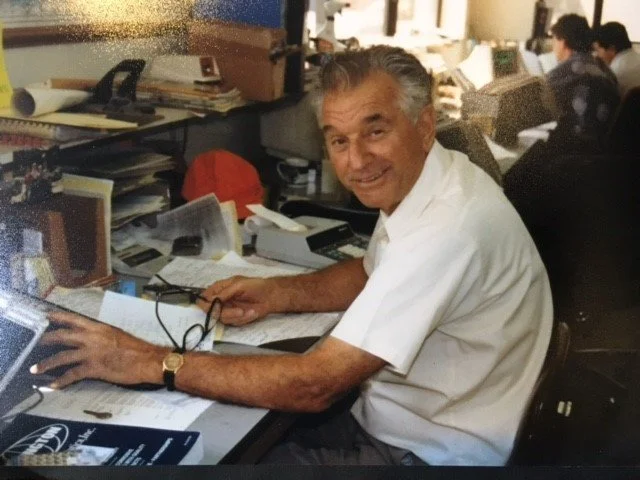 An older man with gray hair smiling at a cluttered office desk with papers, glasses, and a computer, surrounded by other workers in an busy office environment.