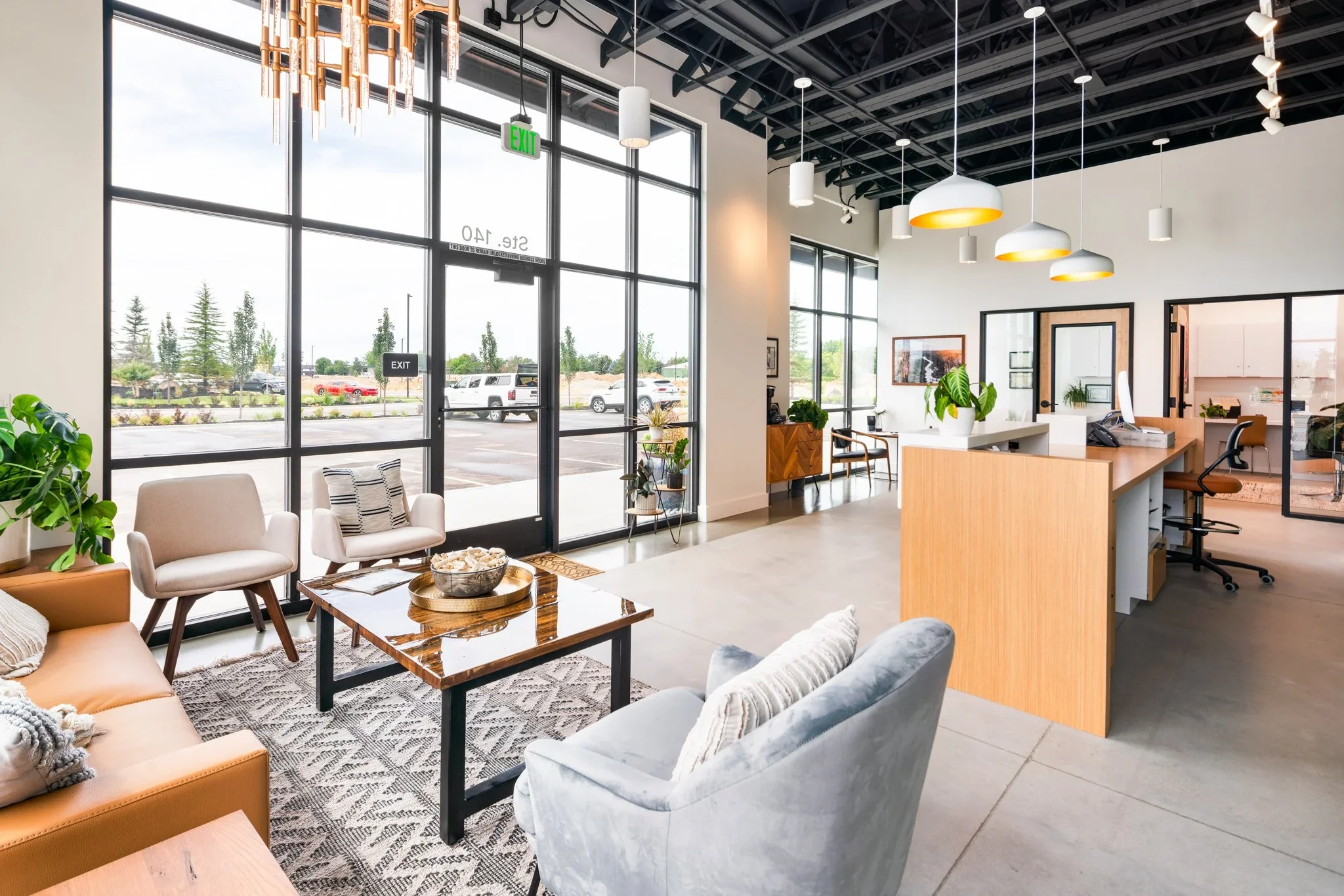 Modern office lobby with large glass windows, seating area with chairs and sofa, a wooden reception desk, potted plants, and pendant lights hanging from a exposed ceiling.