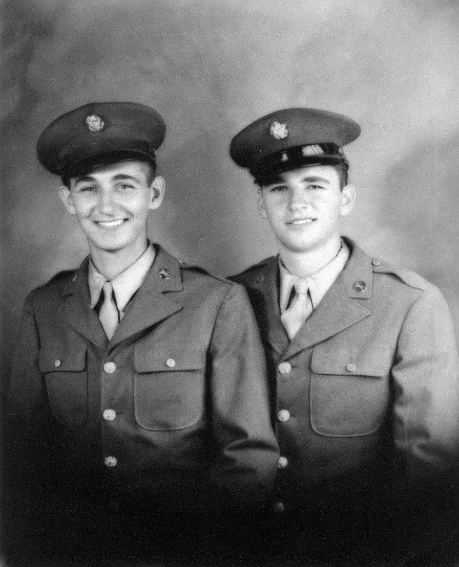 Two young men in vintage military uniforms and hats, standing side by side, smiling in a studio portrait.