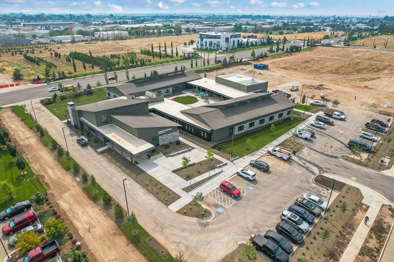 Aerial view of a commercial building with a parking lot and surrounding landscaping, under construction, with empty plots and roads nearby in a developing area.