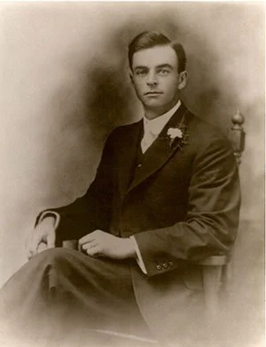 A young man in formal attire, seated on a chair with a neutral expression, in a vintage sepia-toned portrait photograph.