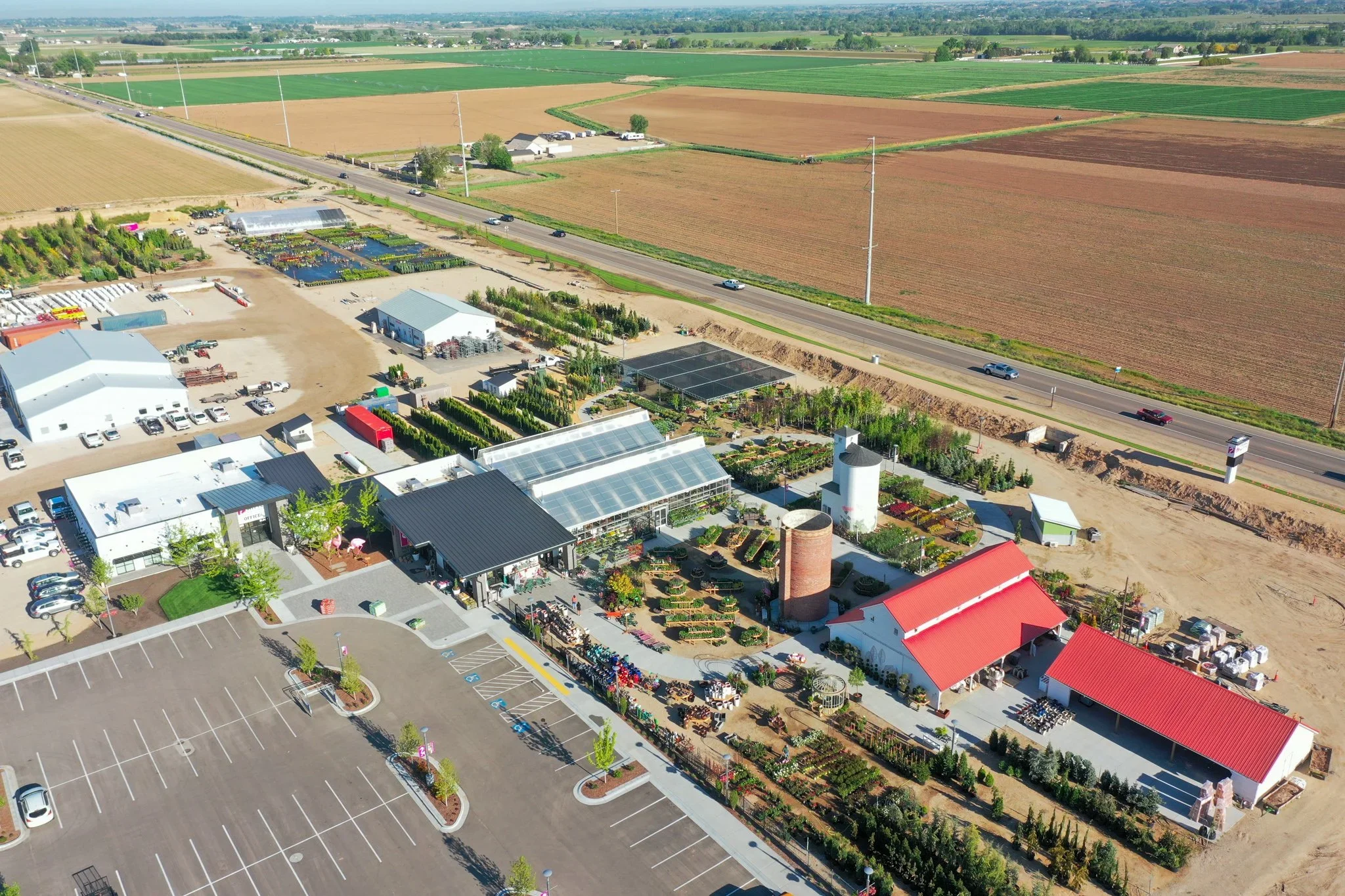 Aerial view of a gardening or nursery business with greenhouses, outdoor plant displays, and a parking lot, surrounded by farmland and highway.