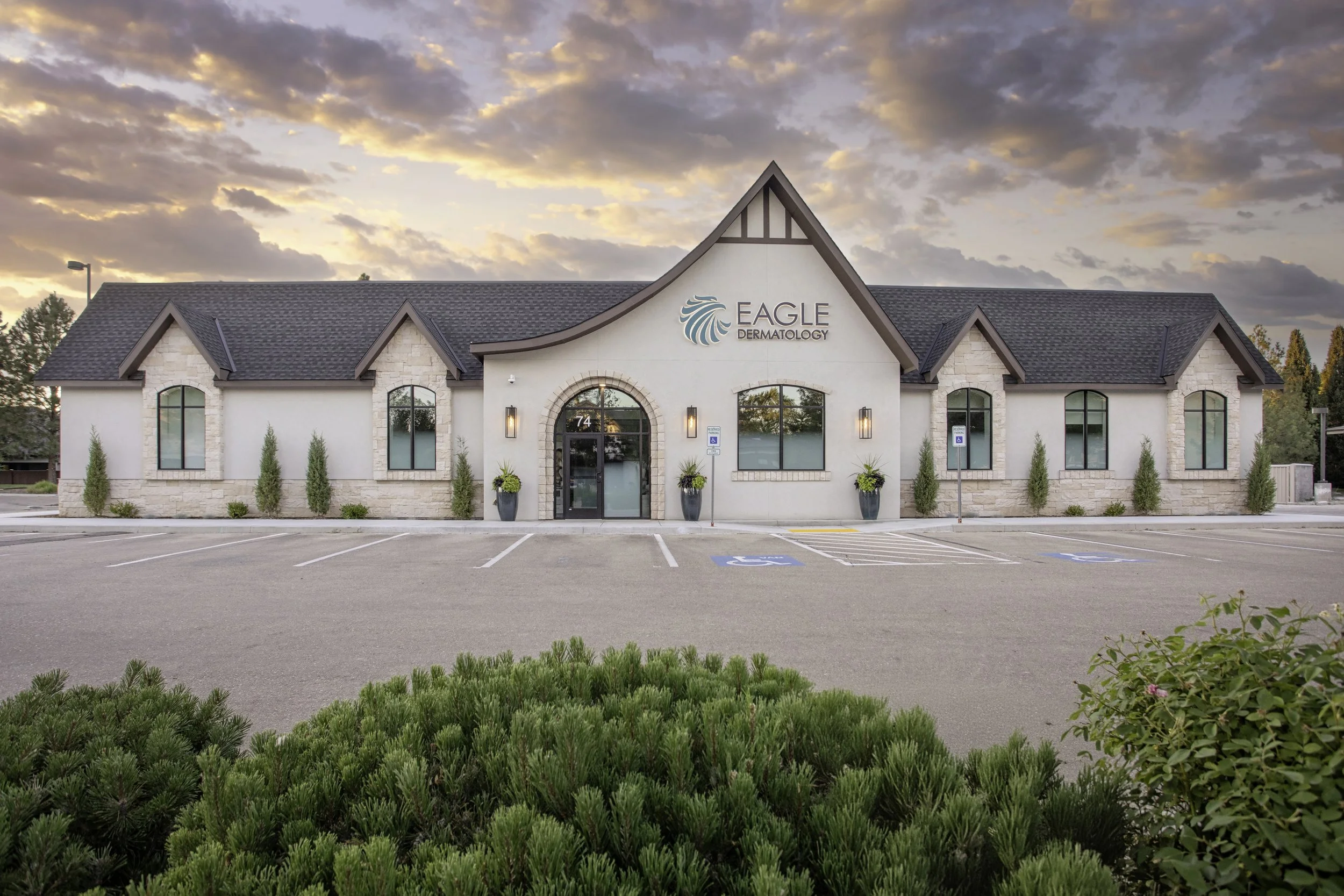 A building with a sign that reads 'Eagle Dermatology,' featuring a stylized eagle logo, beige stone and white exterior, multiple arched windows, and a dark gray roof, with a parking lot in the foreground and a sunset sky with clouds in the background