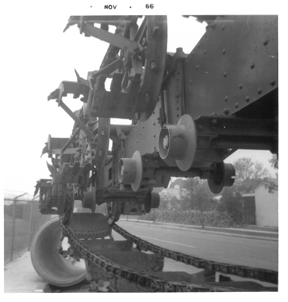 Black and white photo of large machinery, possibly part of a train or industrial equipment, with gears, chains, and rollers, outdoors on a road with a fence and trees in the background.