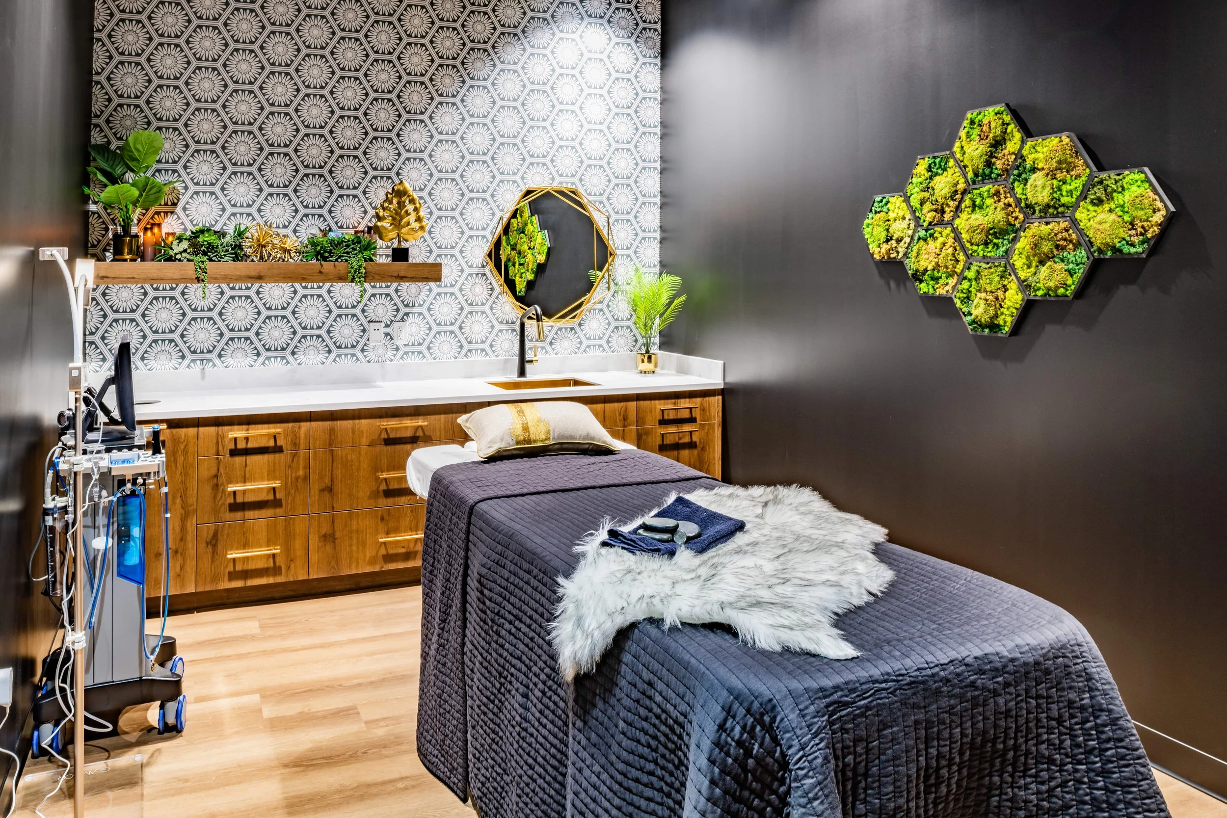 A modern spa treatment room with a massage bed draped in a dark quilt, fluffy white blanket, and a towel with massage tools. The room features a patterned black and white wall, a wooden cabinet with a white countertop, a round mirror, green plants, and decorative wall art with green moss in hexagon-shaped planters.