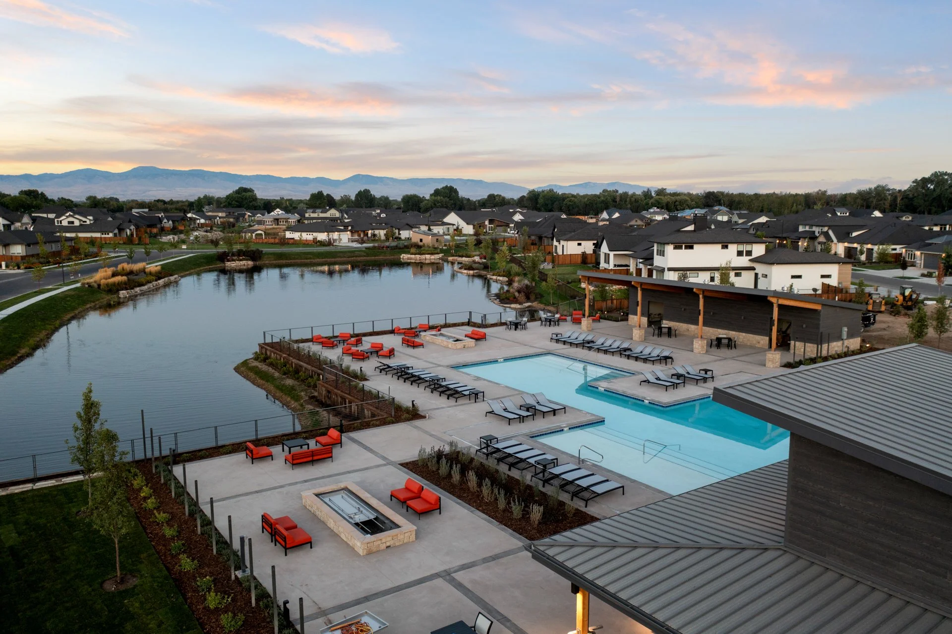 A community swimming pool area with lounge chairs and fire pits, surrounded by a small lake and residential houses under a partly cloudy sky, with mountains in the background.
