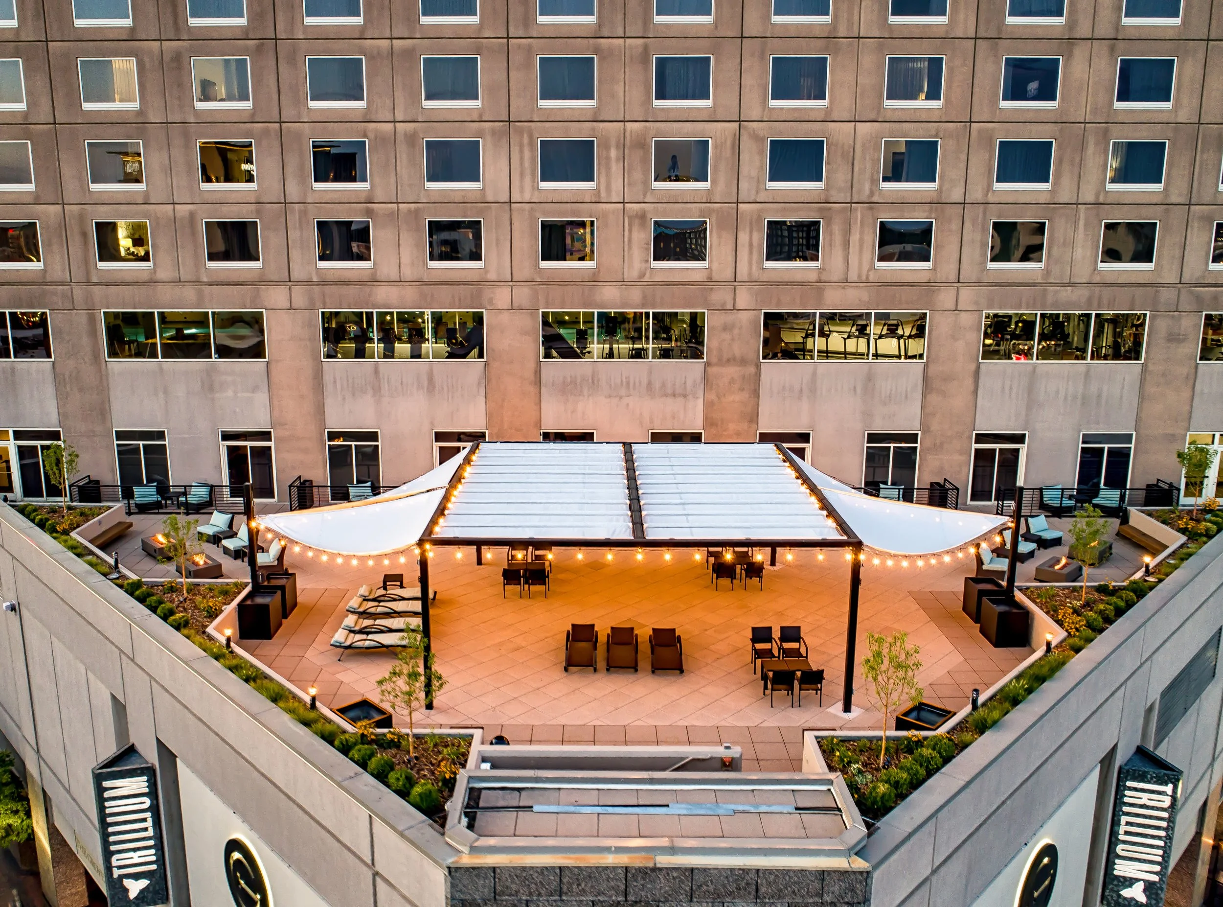 A rooftop patio at sunset featuring string lights, outdoor tables and chairs, sun loungers, and planters with greenery, with a tall building in the background.