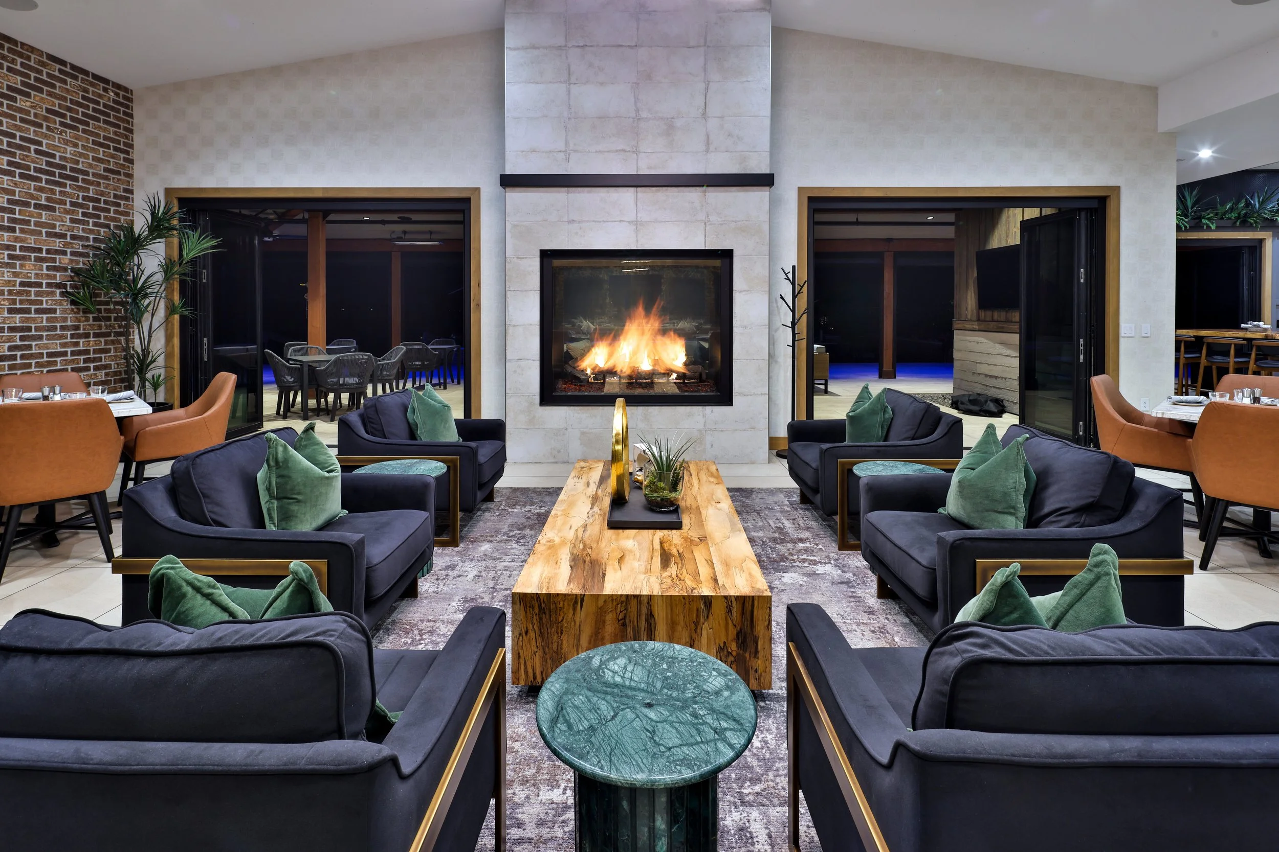 Modern hotel lobby with black sofas, green pillows, a wooden coffee table, a candle centerpiece, a fireplace, and sliding glass doors leading to outdoor seating.