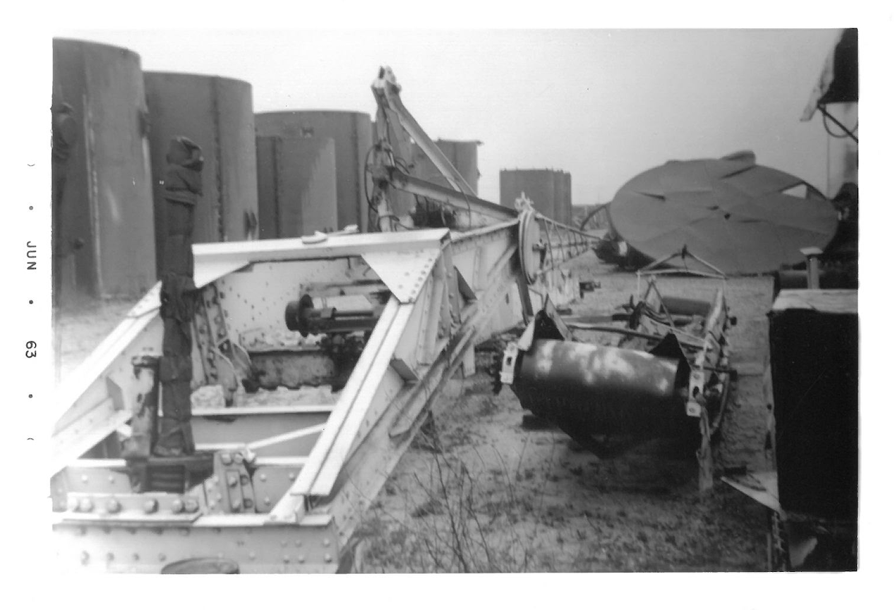 Black and white photo of a fallen military crane and damaged equipment on a desert-like landscape, with large cylindrical structures and a tent in the background.