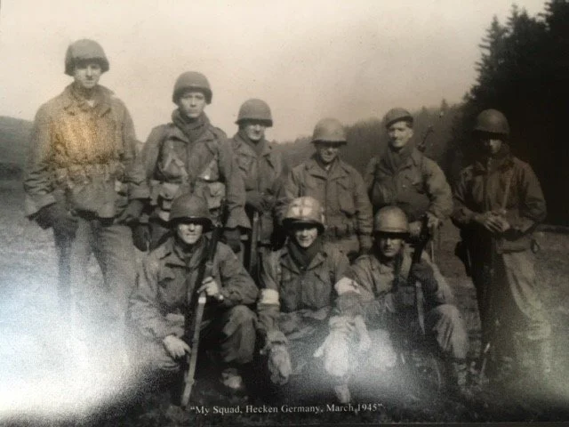 A black-and-white photo of nine soldiers in military uniforms and helmets, some holding rifles, outdoors in a field with trees in the background, taken in March 1945 during World War II.