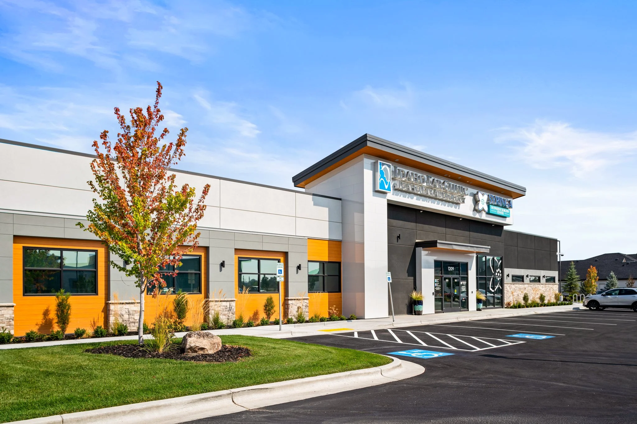 Front view of a modern medical building with a parking lot, including accessible parking spots, a tree with fall foliage, and a clear blue sky.
