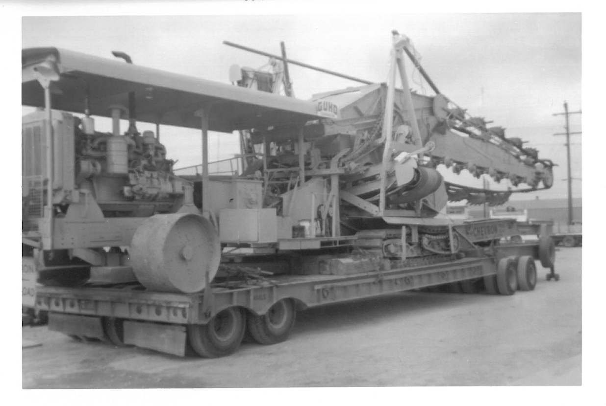 A large construction or industrial machine being transported on a flatbed trailer, with a clear sky and power lines in the background.