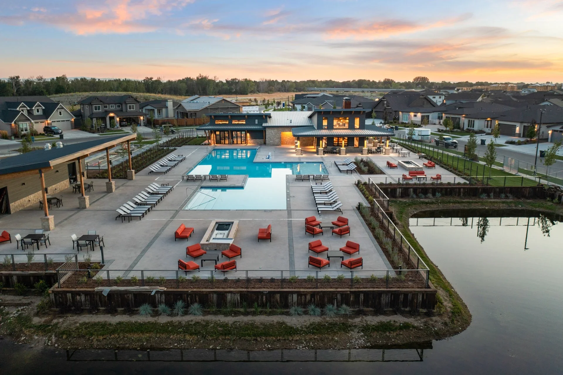 An aerial view of a modern residential community pool area at sunset, featuring a large outdoor pool, several lounge chairs, seating arrangements, a hot tub, and landscaped surroundings, with neighboring houses and a pond nearby.
