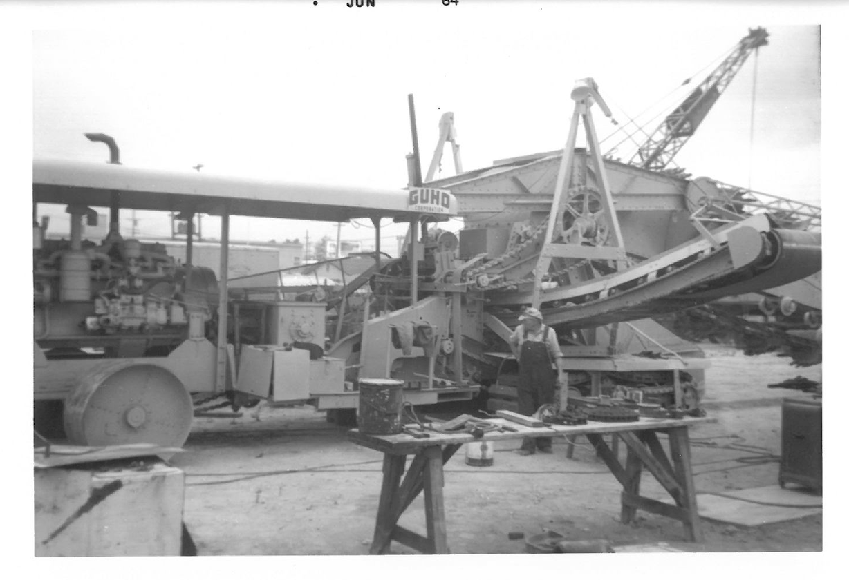 Black and white photograph of an oil drilling rig with a worker standing nearby, surrounded by tools and equipment.