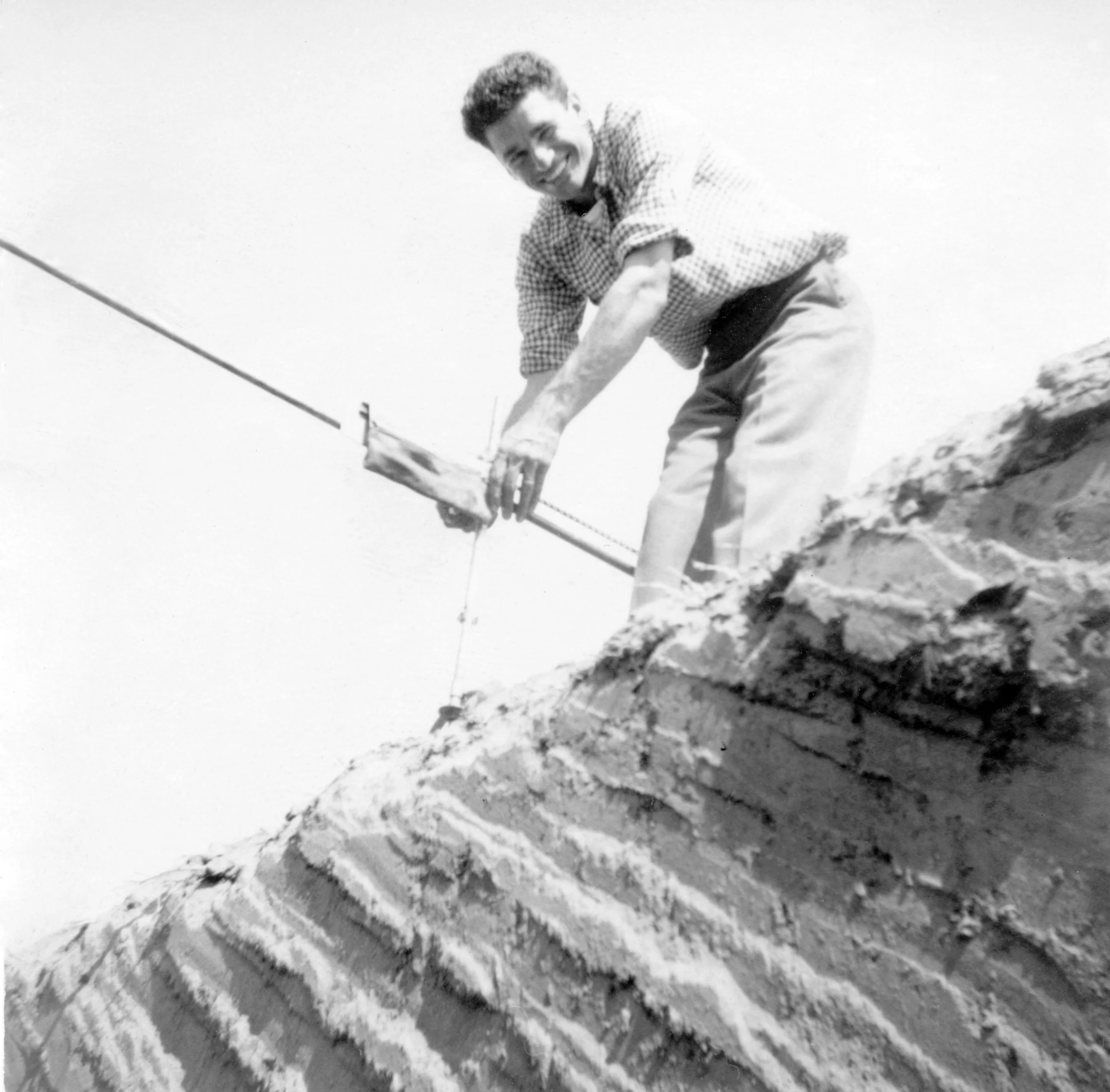 A young man with curly hair smiling while working on a construction site, crouched on a brick wall, using a tool with a long pole.