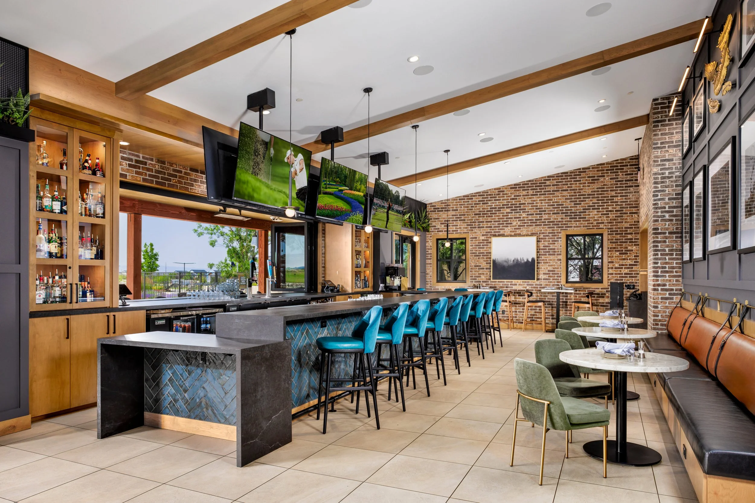 Interior of a modern bar with a long counter, blue barstools, wall-mounted televisions, and a seating area with marble tables and green chairs, featuring brick walls and framed artwork.