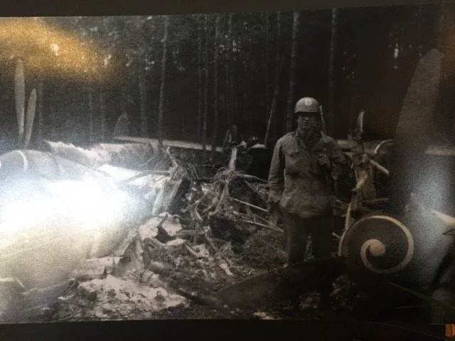A firefighter in uniform and helmet standing amidst the wreckage of a destroyed vehicle in a forested area at night.