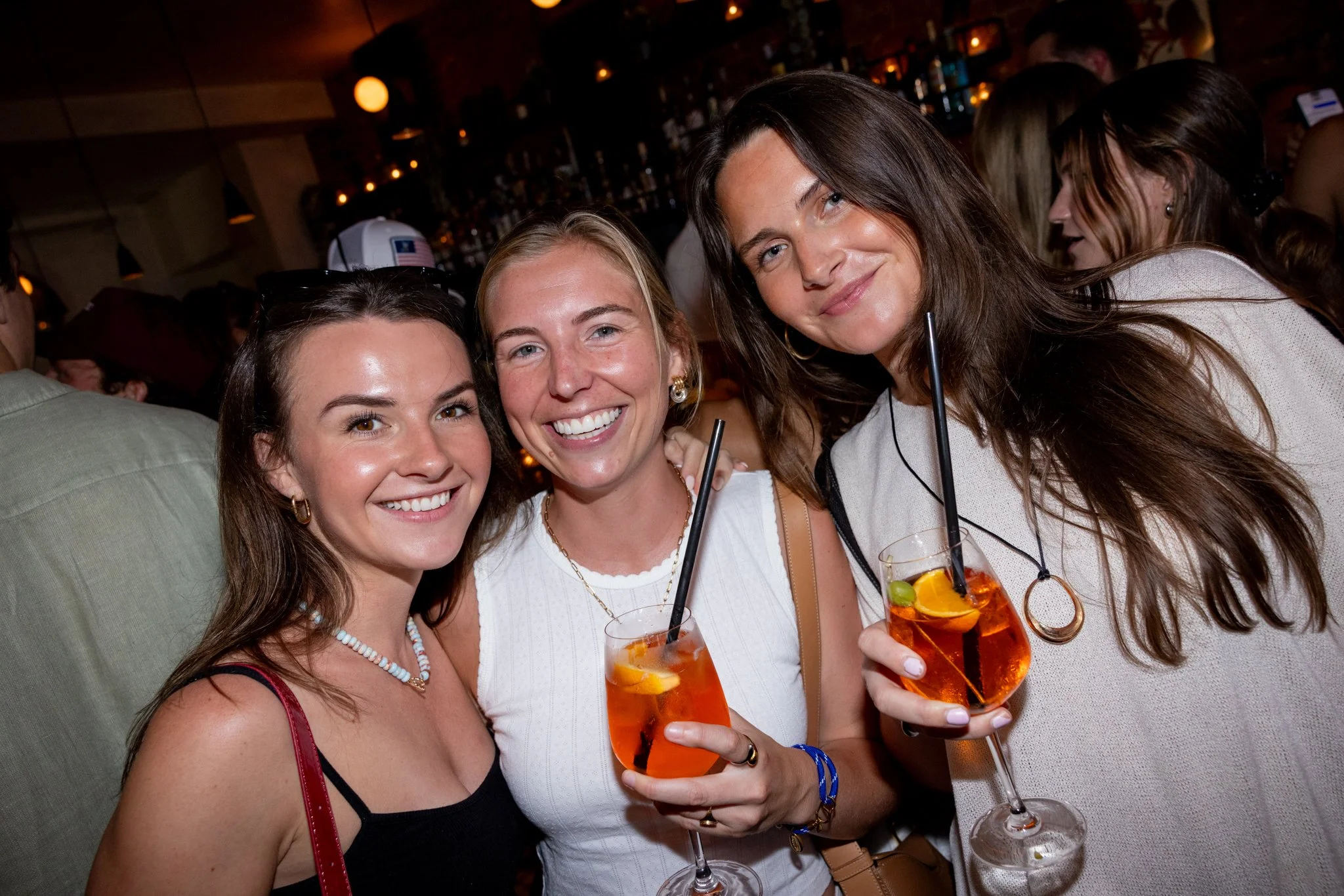 Three women smiling and holding cocktails in a bar or restaurant setting with other people in the background.