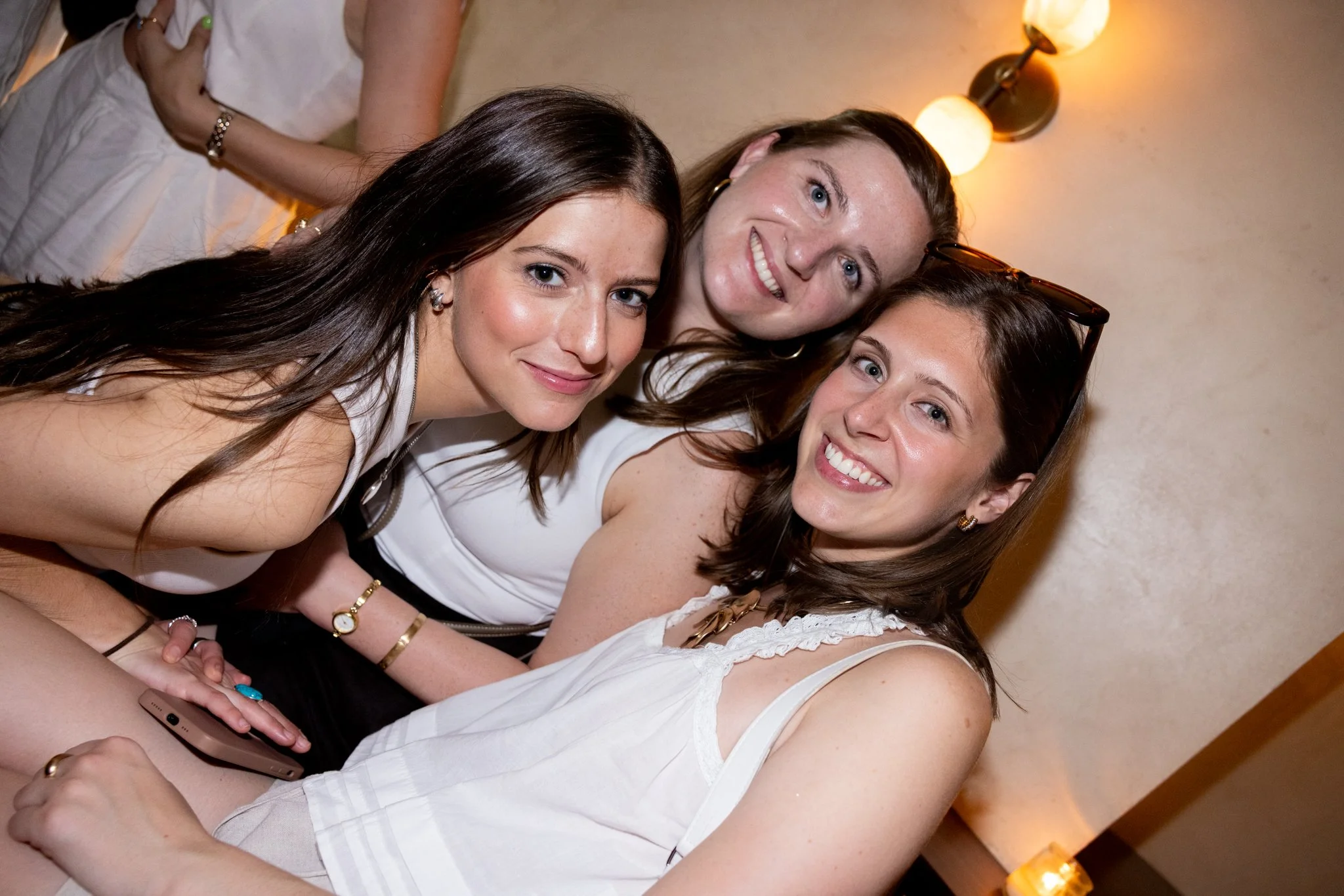 Three young women smiling and leaning close together at an indoor gathering.