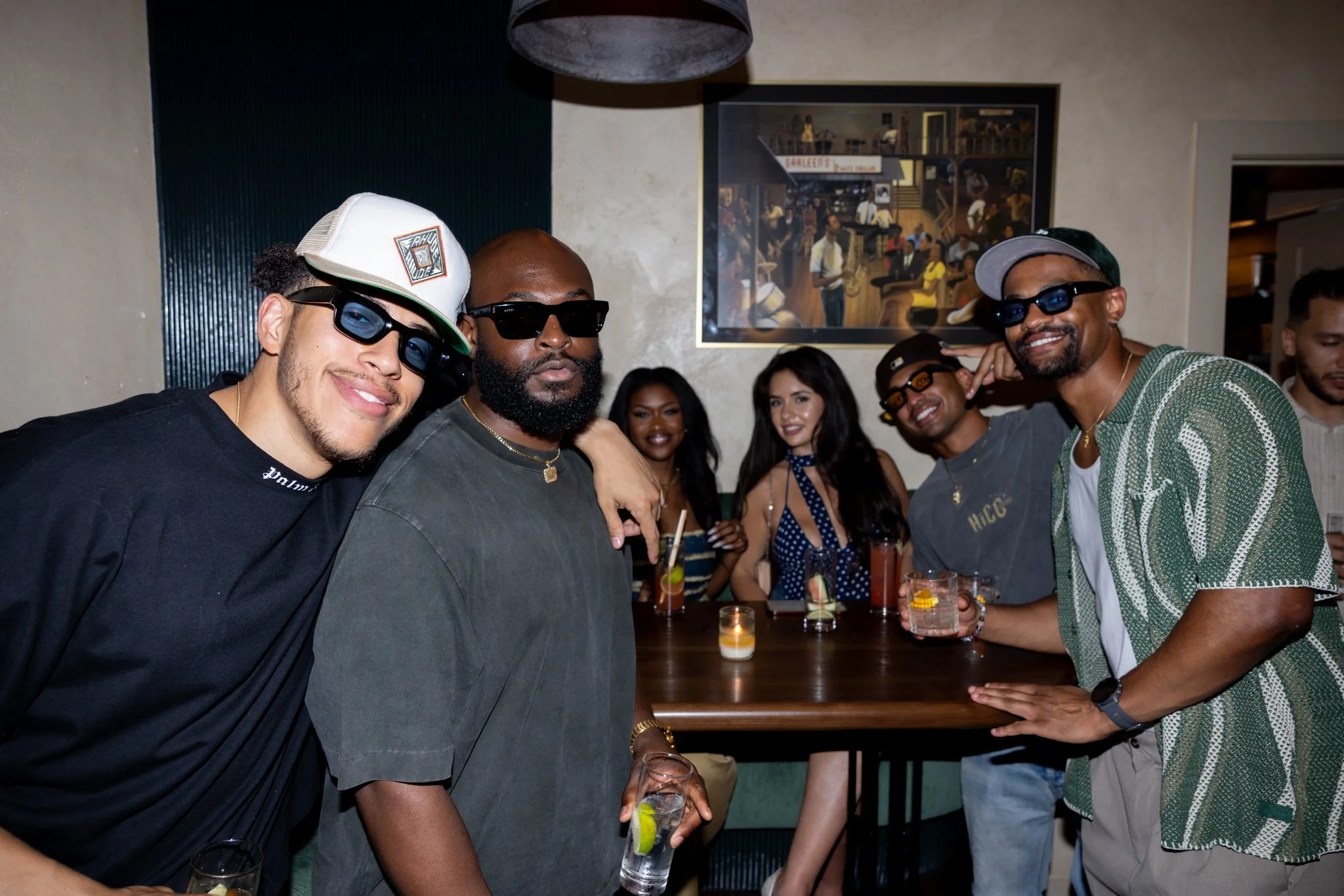 Group of friends smiling and enjoying drinks at a bar or lounge, with some holding glasses and others posing for the camera, in a casual indoor setting.