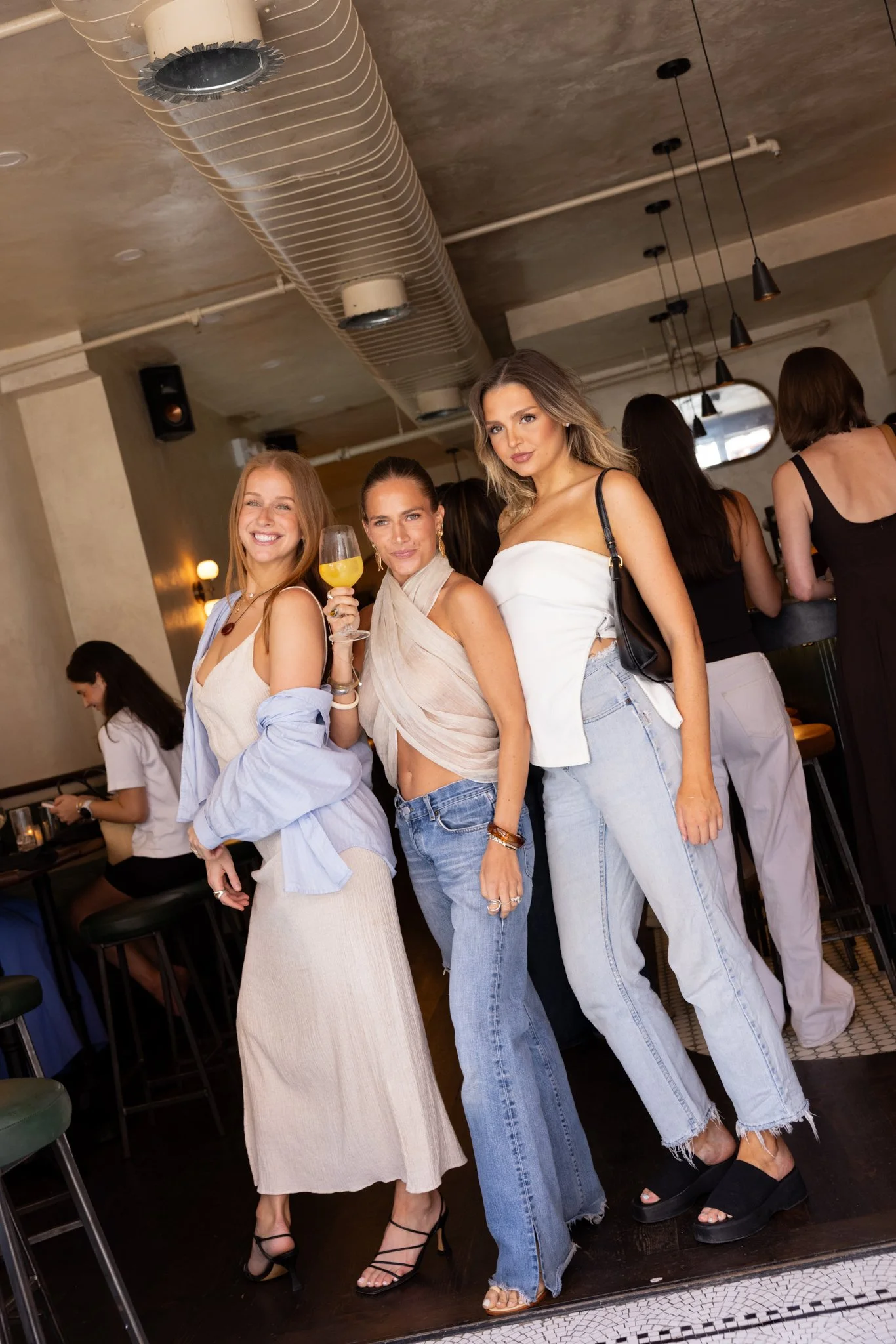 Three women enjoying a night out at a bar or restaurant, with some women in the background, the woman in the middle is holding a glass of orange juice, all dressed casually and stylishly.