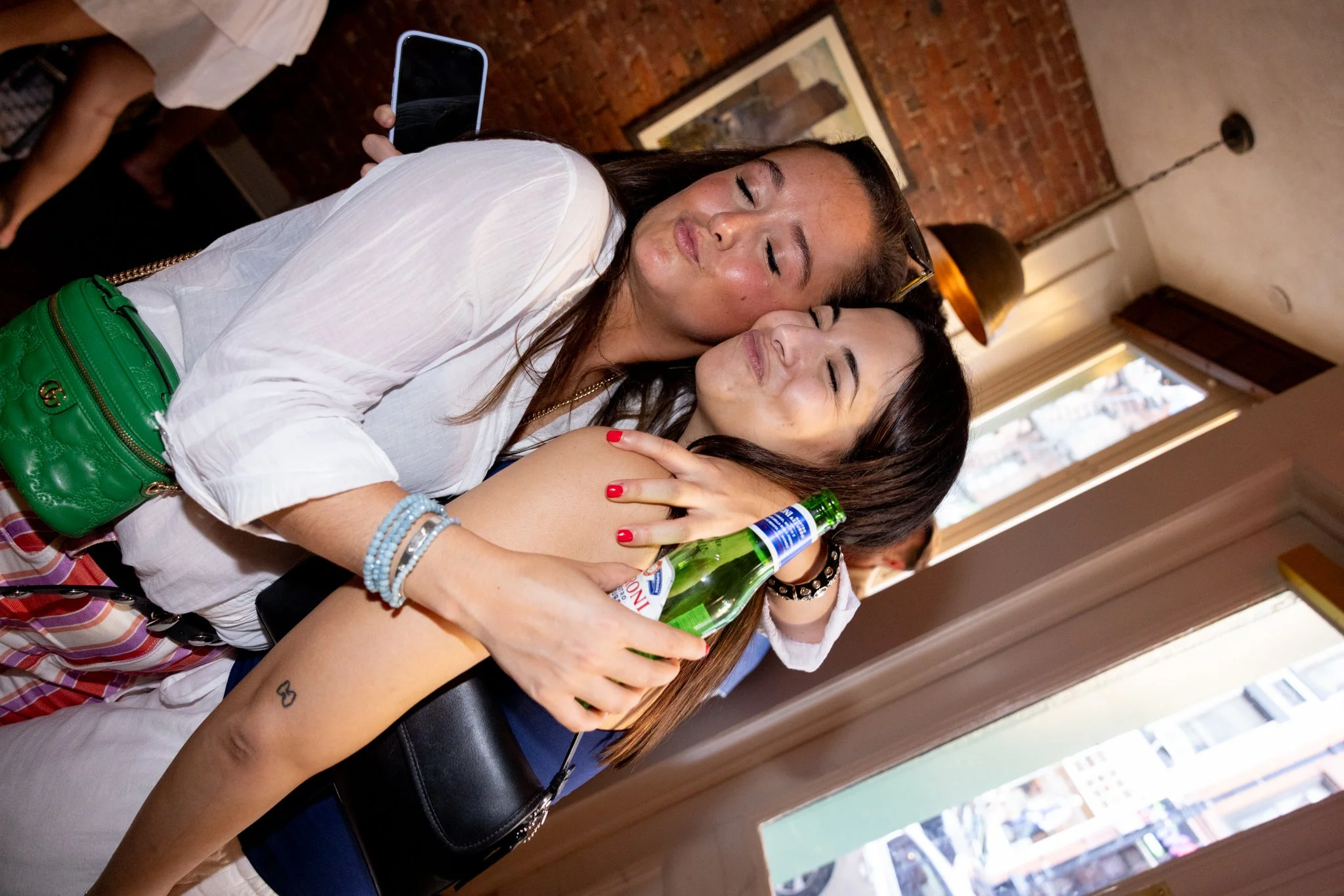 Two women hugging and celebrating with a bottle of beer in front of exposed brick wall and large window.