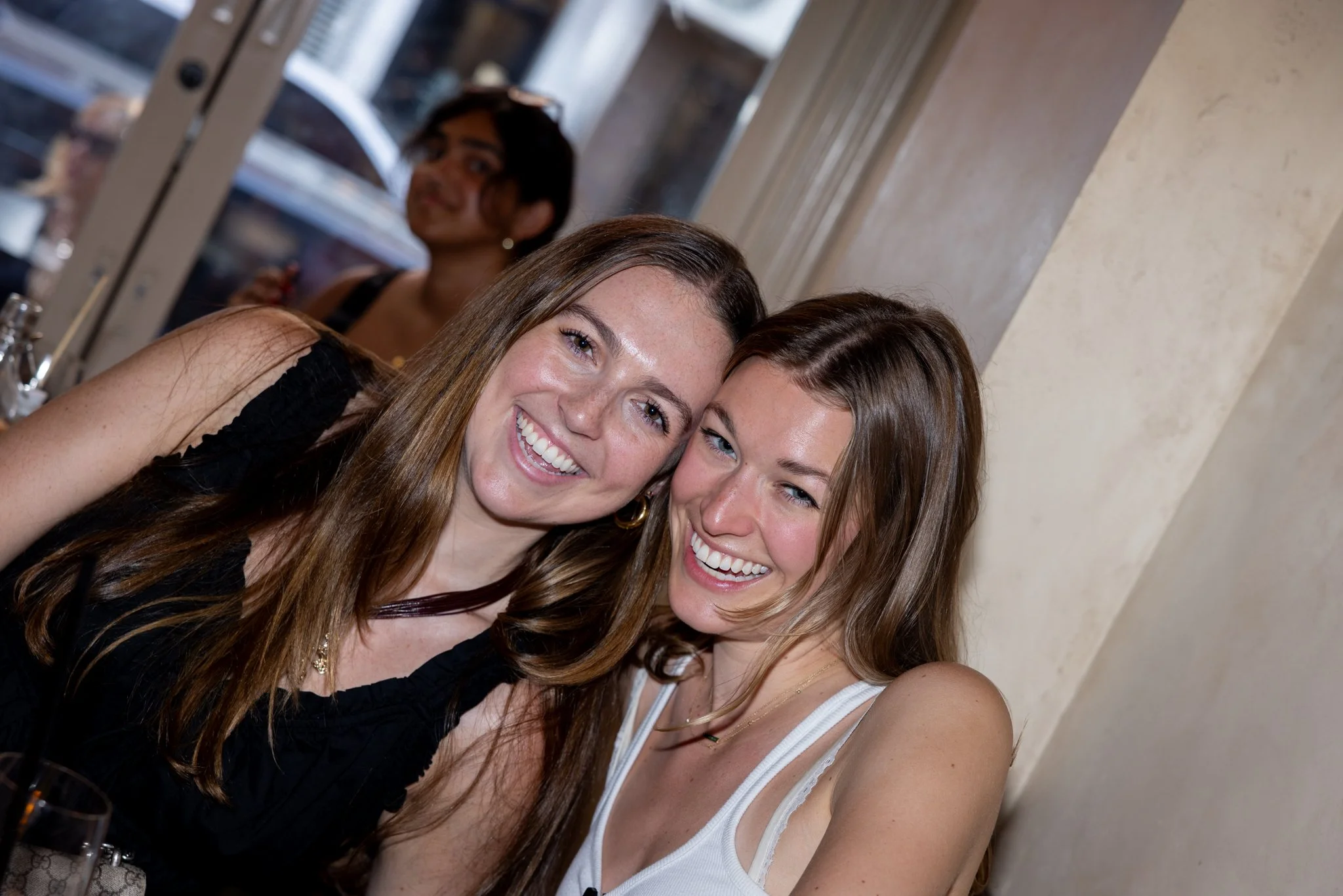 Two young women smiling and leaning their heads together at a social gathering in a restaurant or cafe, with a third person in the background near a window.